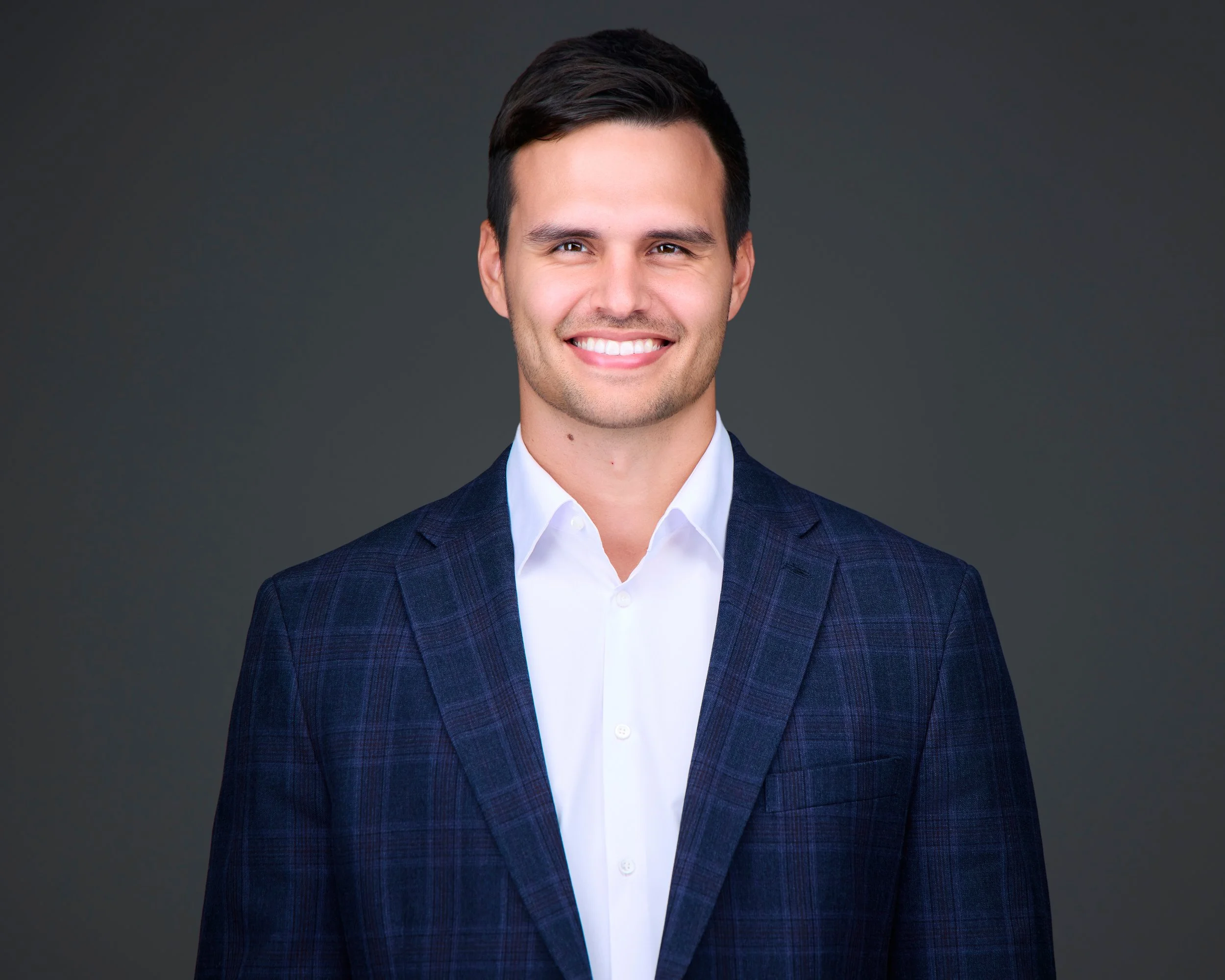 A smiling young man with dark hair, dressed in a white shirt and a dark blue plaid suit jacket, against a plain dark background.