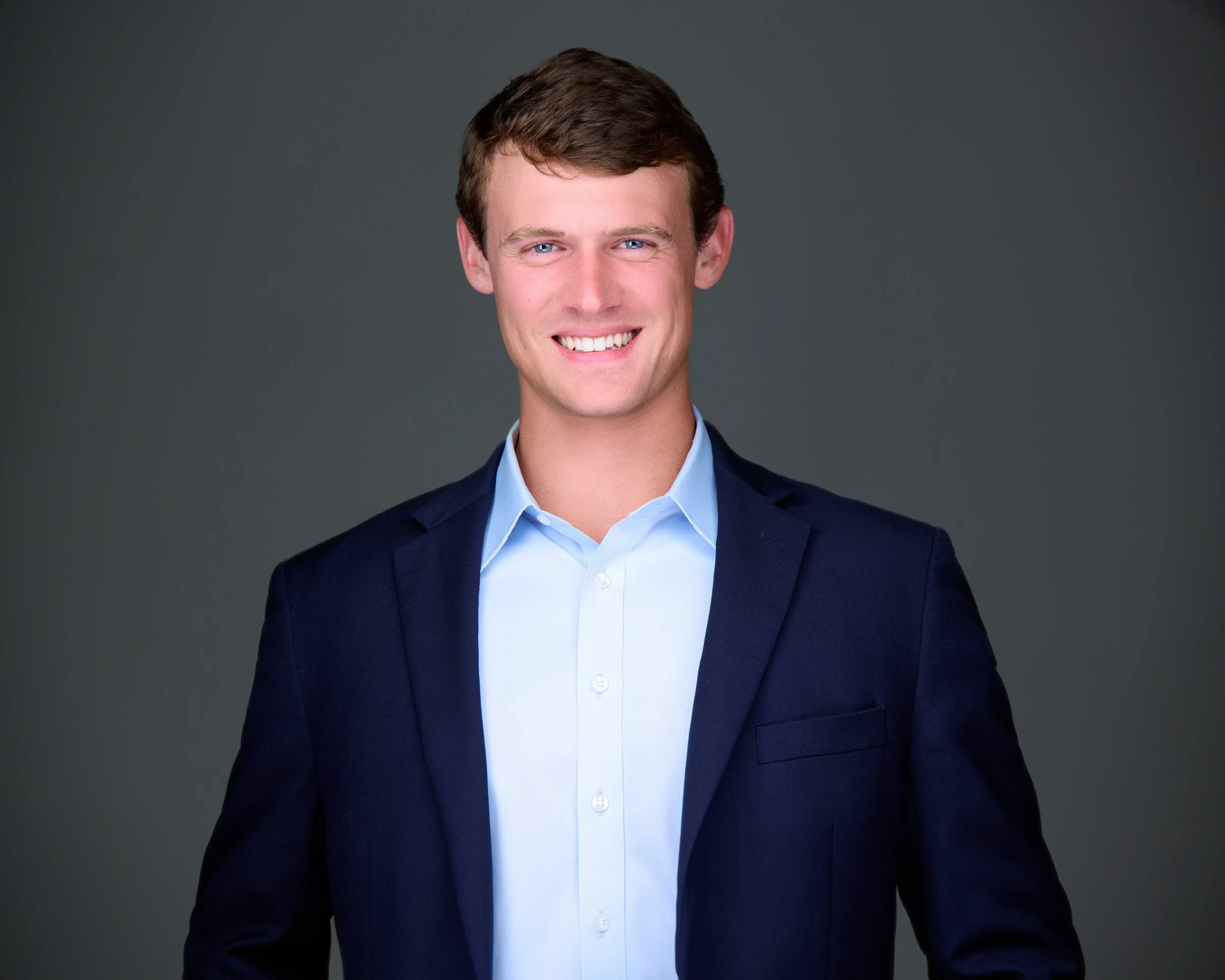 A young man with brown hair and blue eyes, wearing a blue suit jacket over a light blue dress shirt, smiling against a dark gray background.