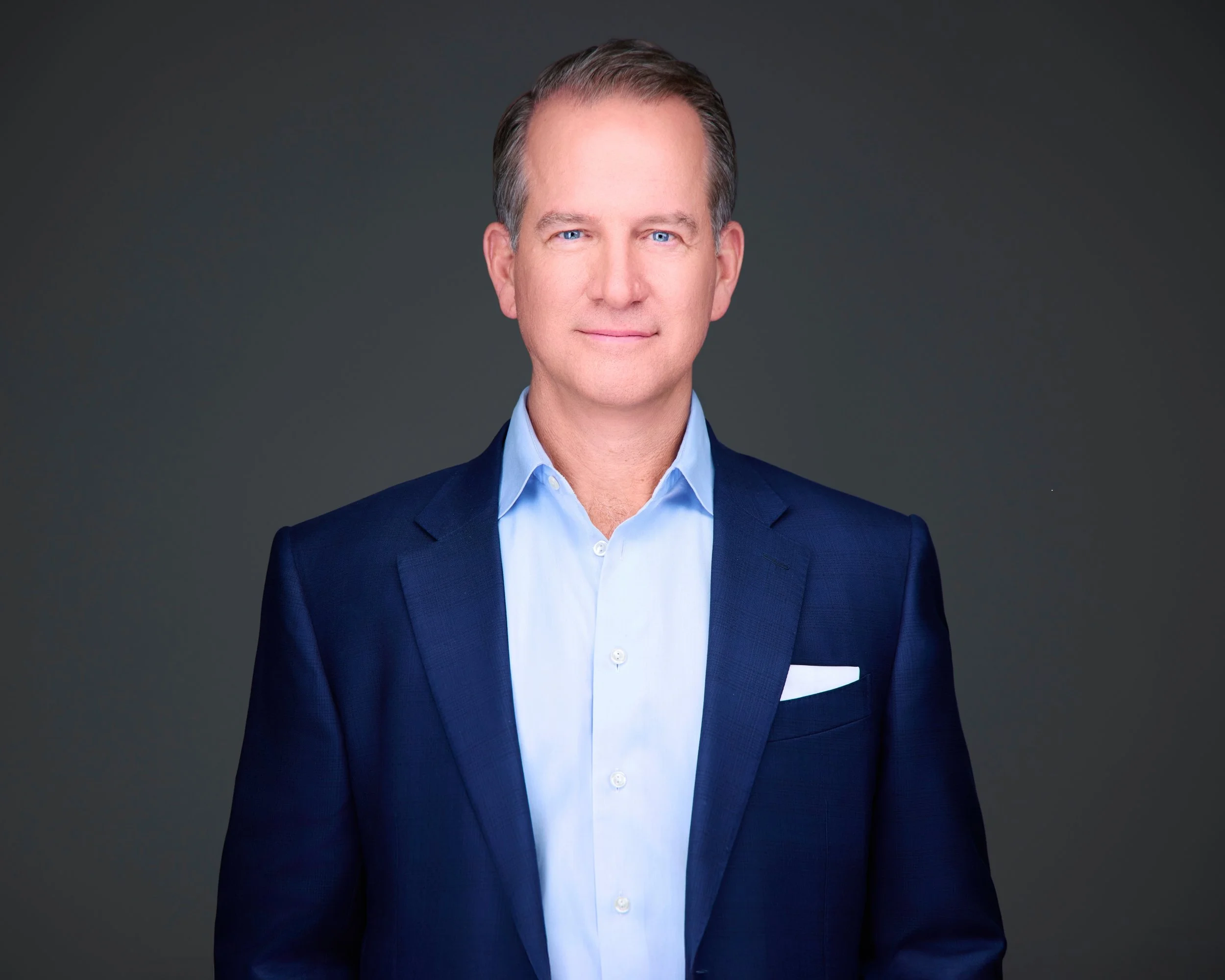 Professional headshot of a man with light skin, short brown hair, wearing a navy blazer, light blue dress shirt, and a white pocket square, standing against a plain dark background.