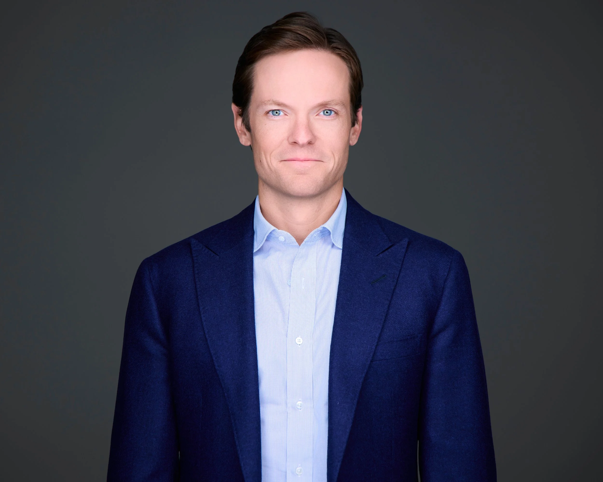 A professional man with light skin, blue eyes, and brown hair styled back, wearing a navy blazer over a light blue button-up shirt, standing against a plain gray background.