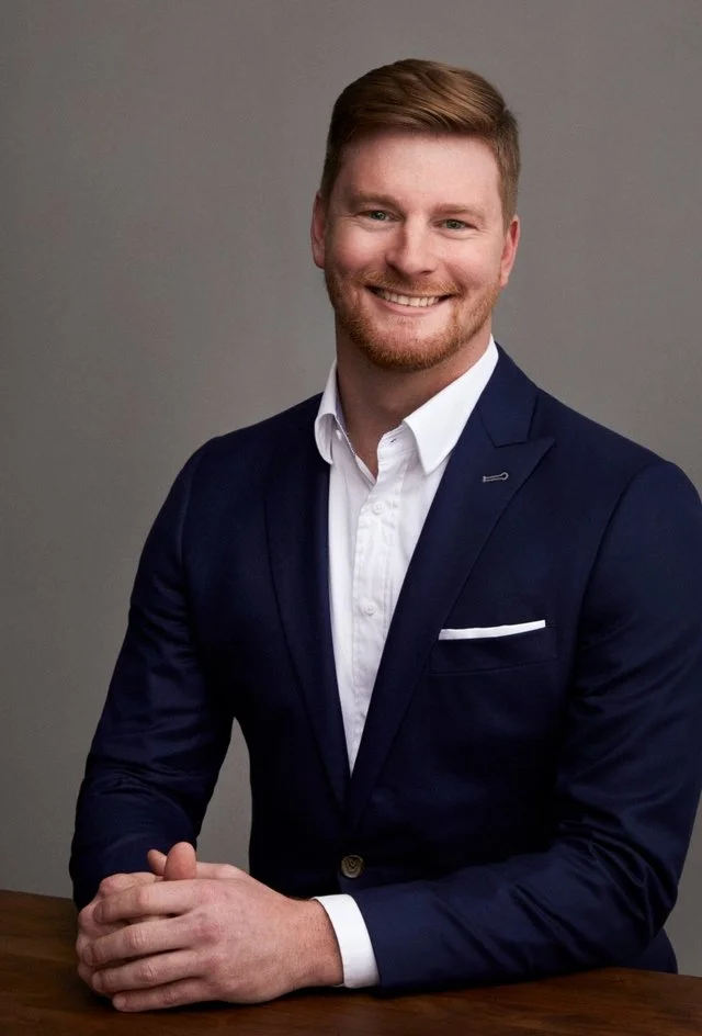 A young man with reddish hair and beard in a navy blazer and white shirt, smiling, seated at a table against a plain gray background.