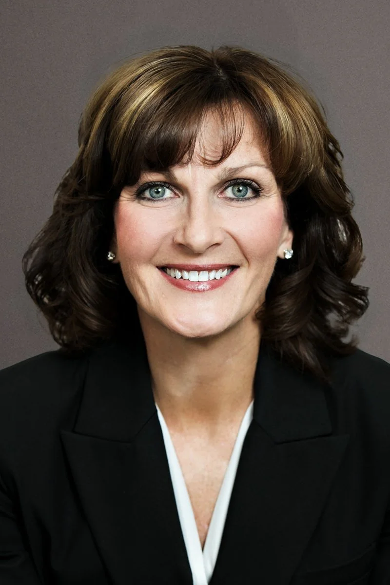 A professional woman with shoulder-length brown hair and blue eyes, smiling and wearing a black blazer and pearl earrings.