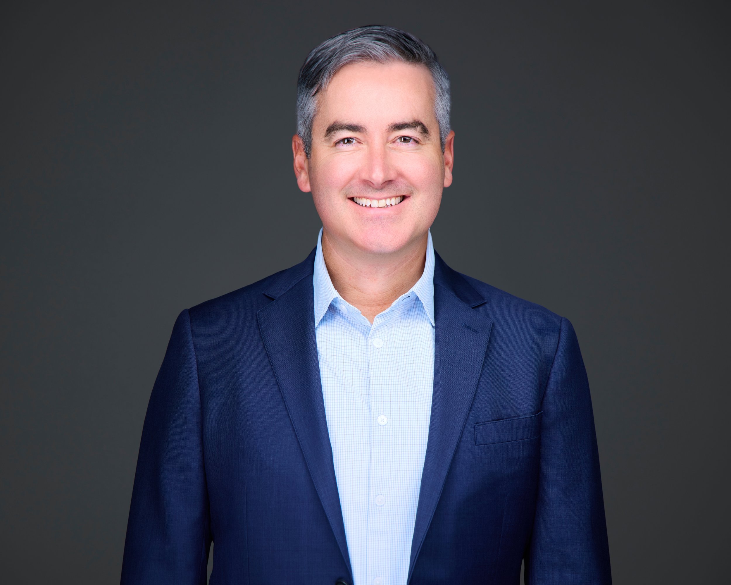Portrait of a smiling man wearing a navy blue blazer and light blue shirt against a dark gray background.