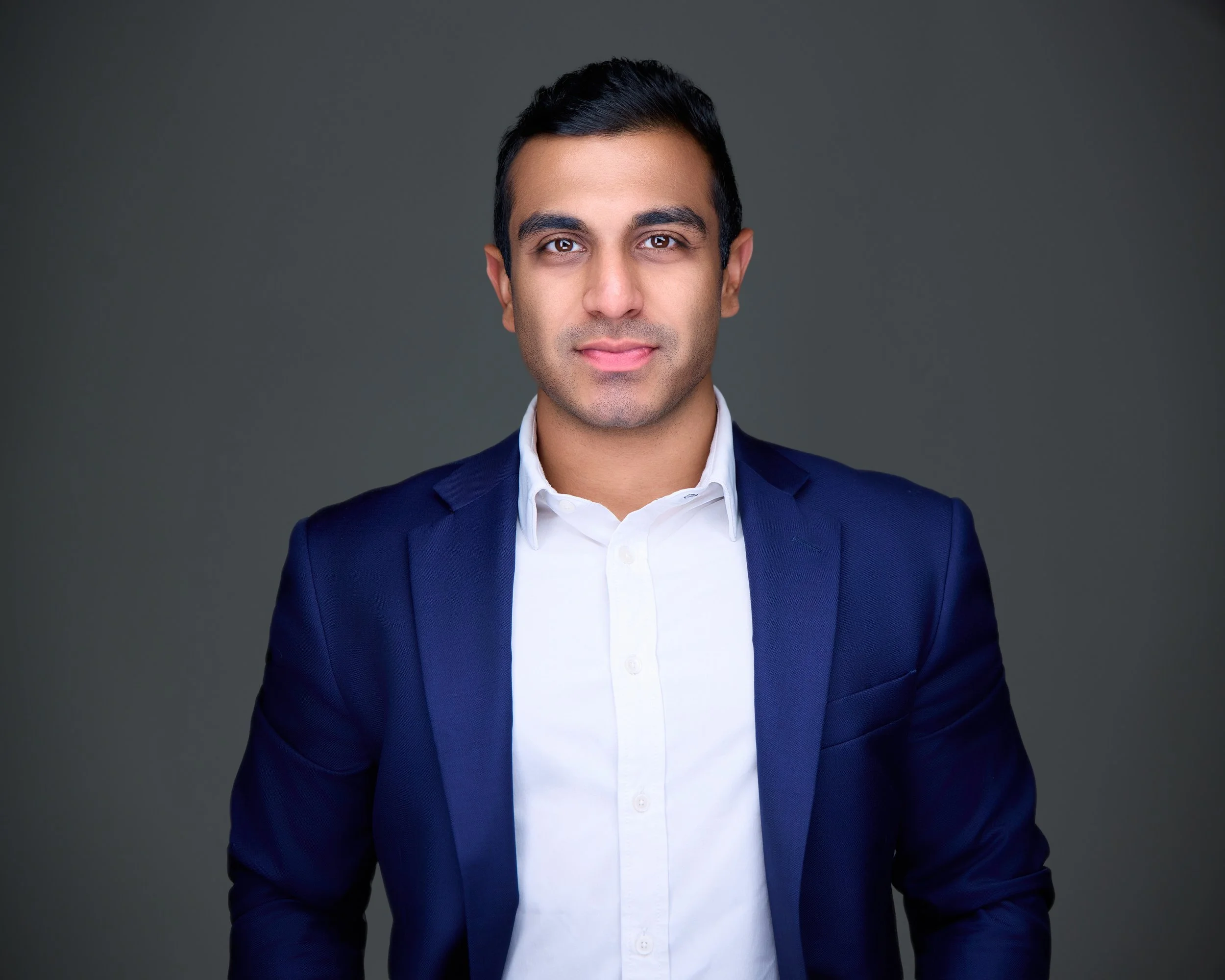Portrait of a young man in a dark blue blazer and white shirt, standing against a gray background.