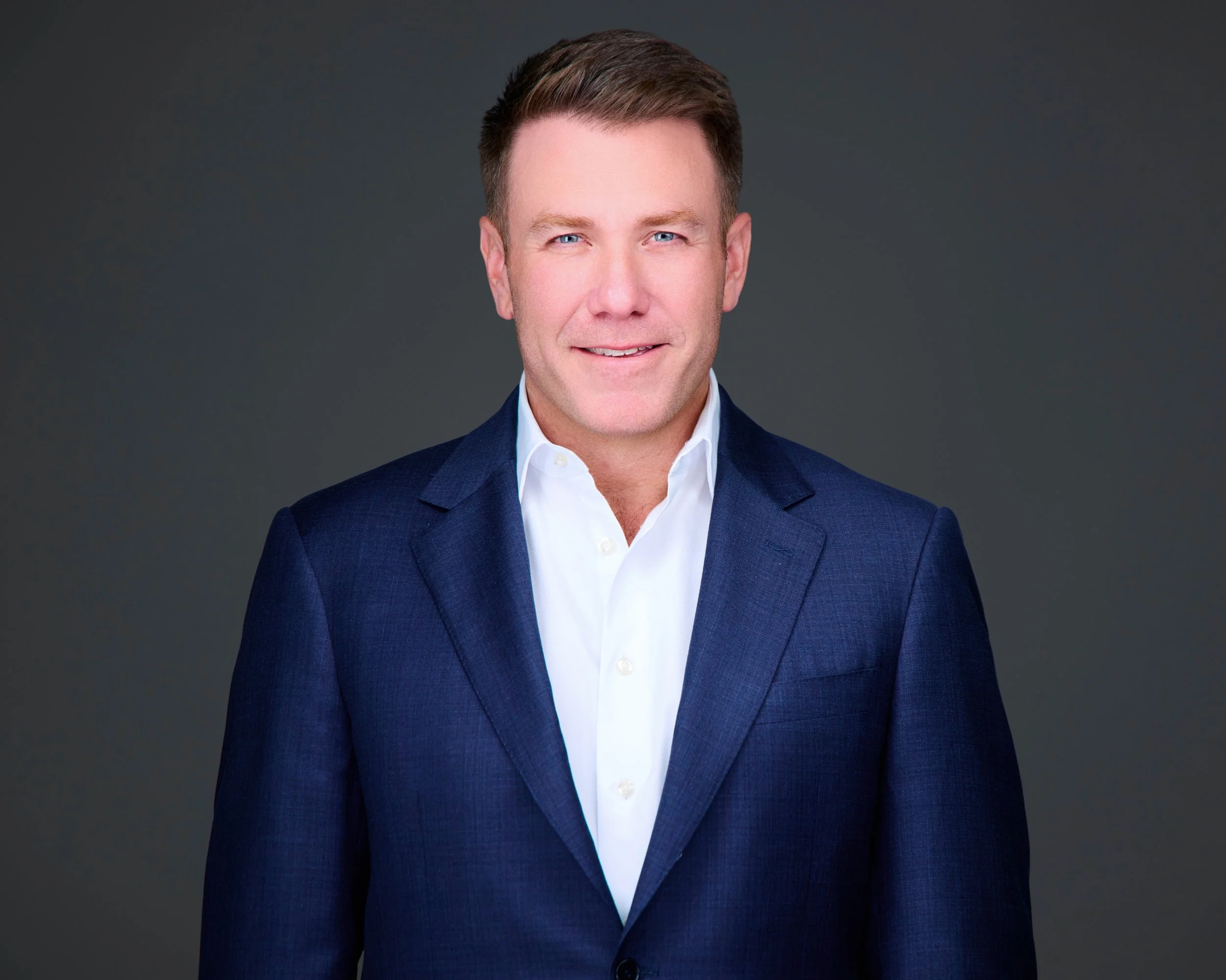 A man with short brown hair wearing a navy blue suit jacket and white dress shirt smiling at the camera against a dark gray background.