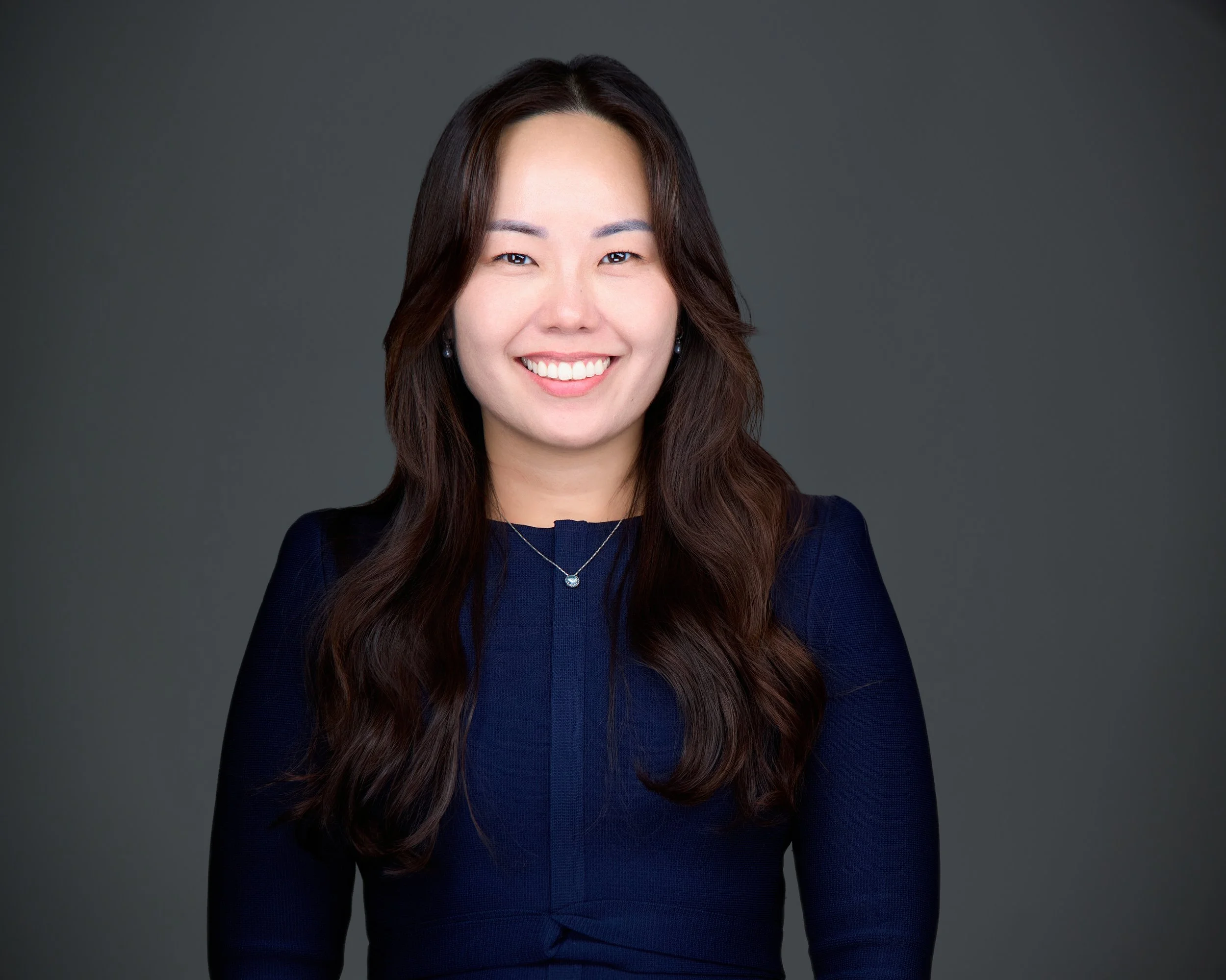 A professional woman with long brown hair, smiling, wearing a navy blue dress and jewelry, against a gray background.