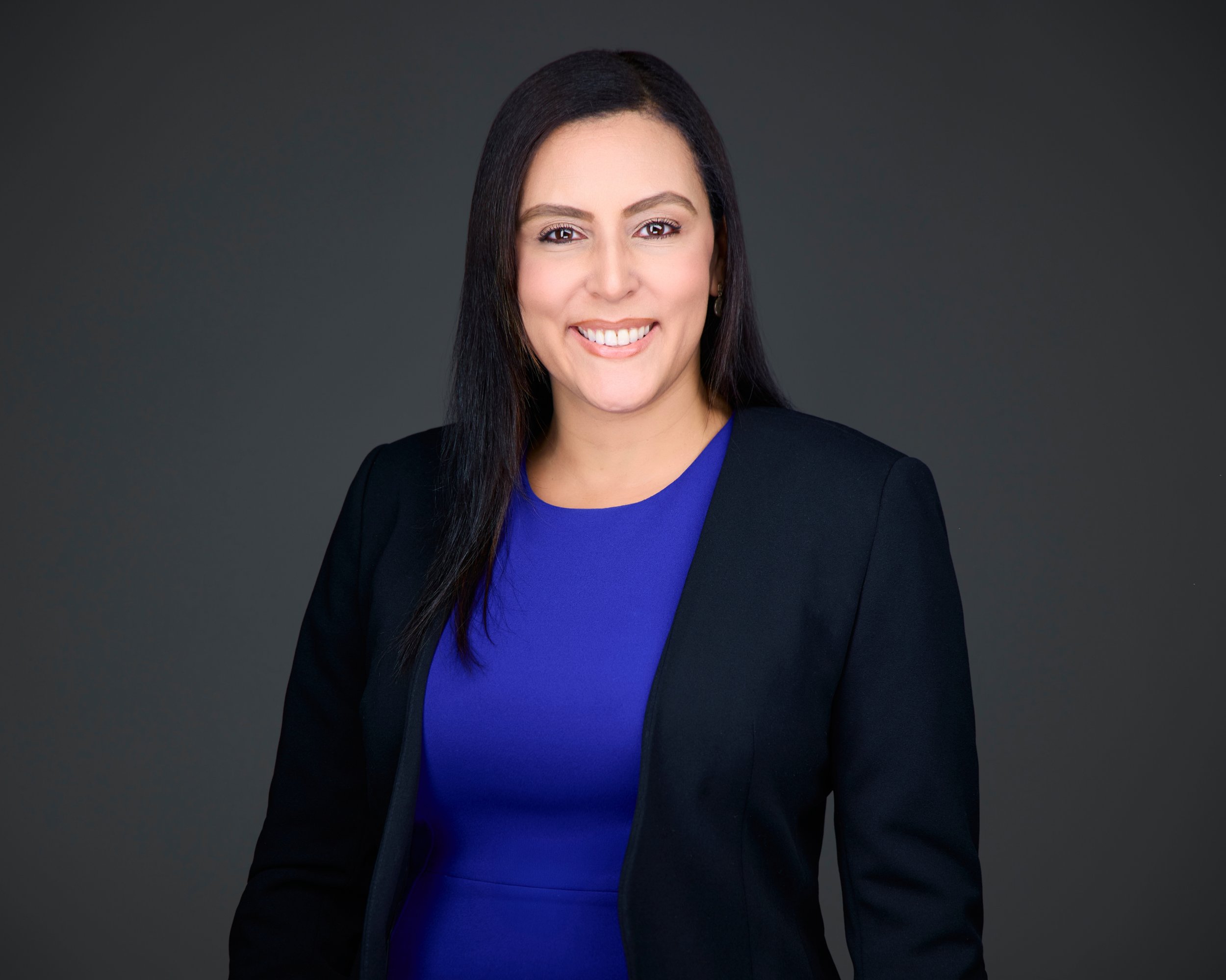 Professional woman with dark hair wearing a black blazer and blue top, smiling against a dark background.