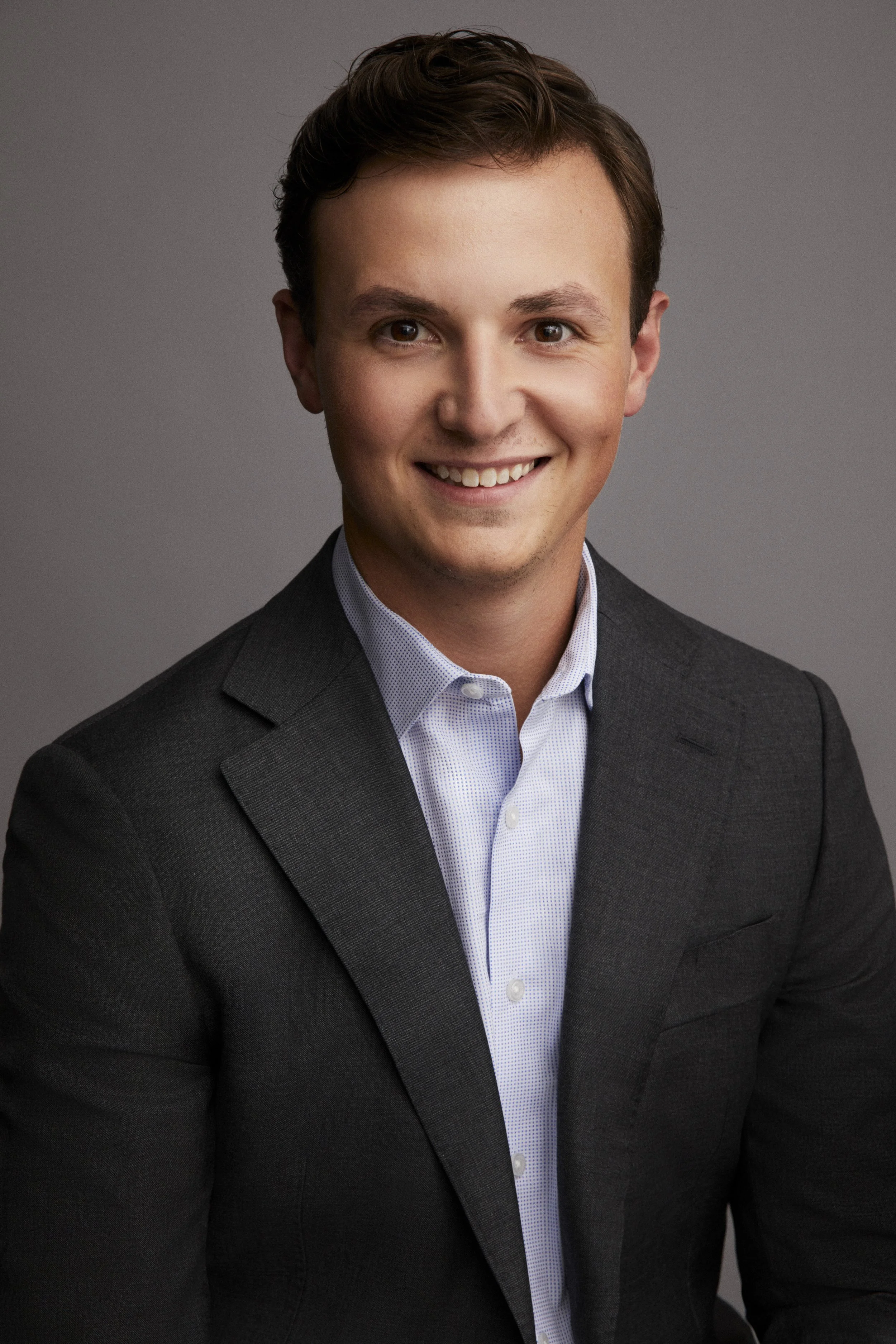 A portrait of a young man with short brown hair, smiling, wearing a dark suit jacket over a light collared shirt, against a plain gray background.