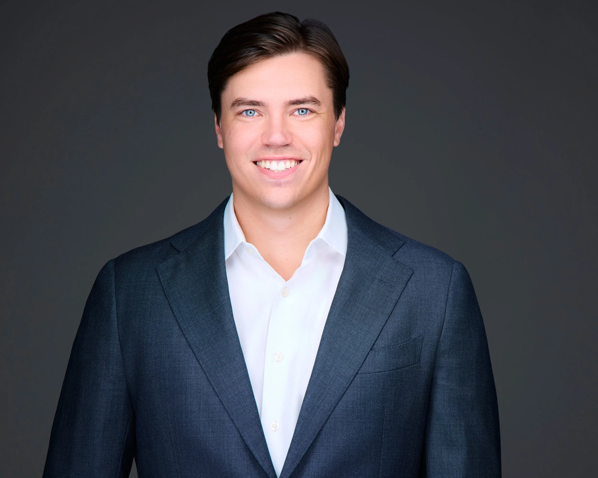 A young man with dark hair and blue eyes in a white dress shirt and navy suit posing against a gray background.