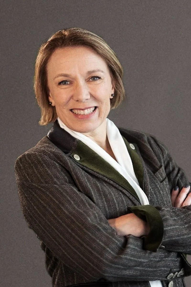 A smiling woman with short brown hair wearing a dark pinstripe blazer over a white shirt, posing with arms crossed against a gray background.