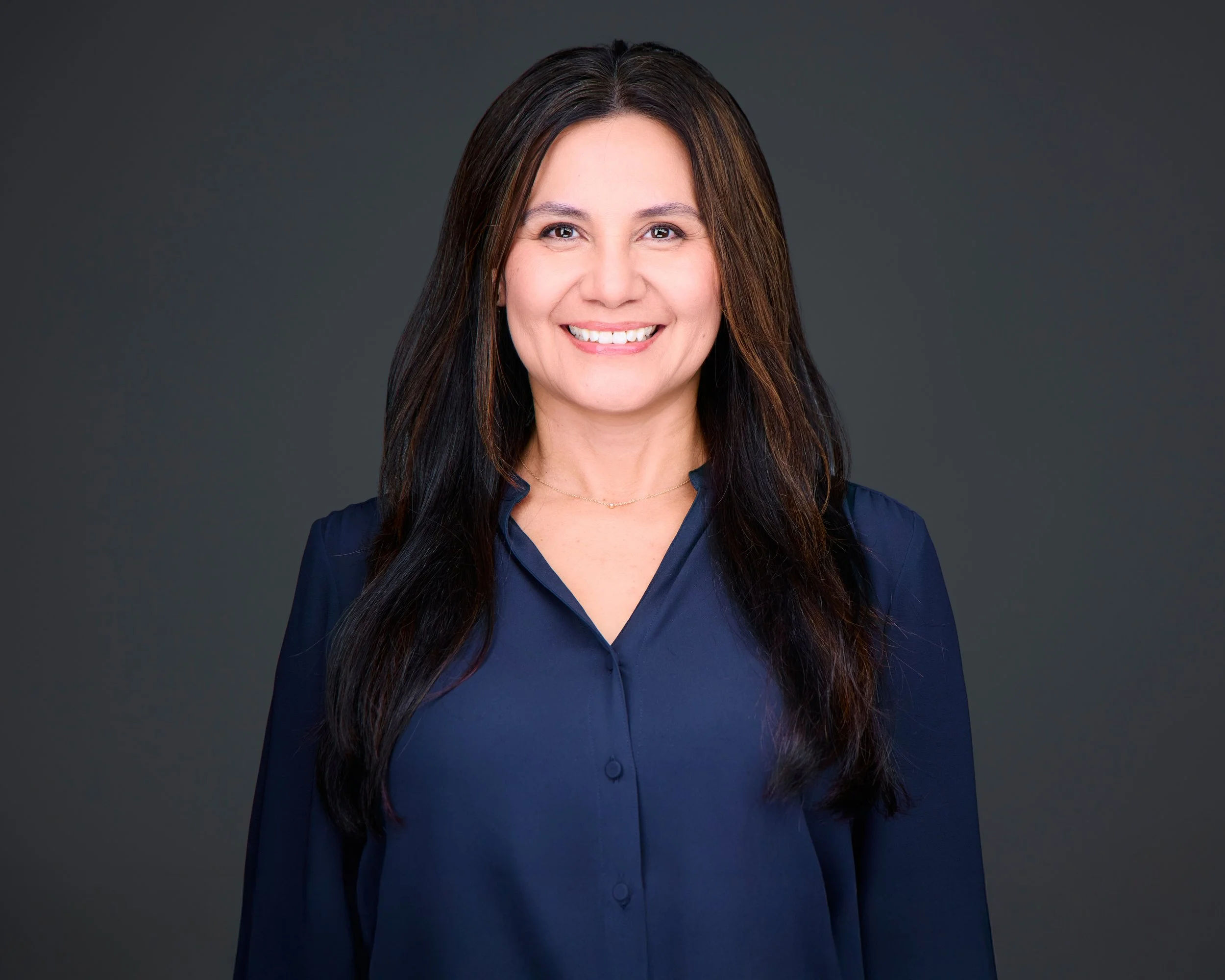 A woman with long dark hair wearing a navy blue blouse, smiling at the camera against a dark gray background.