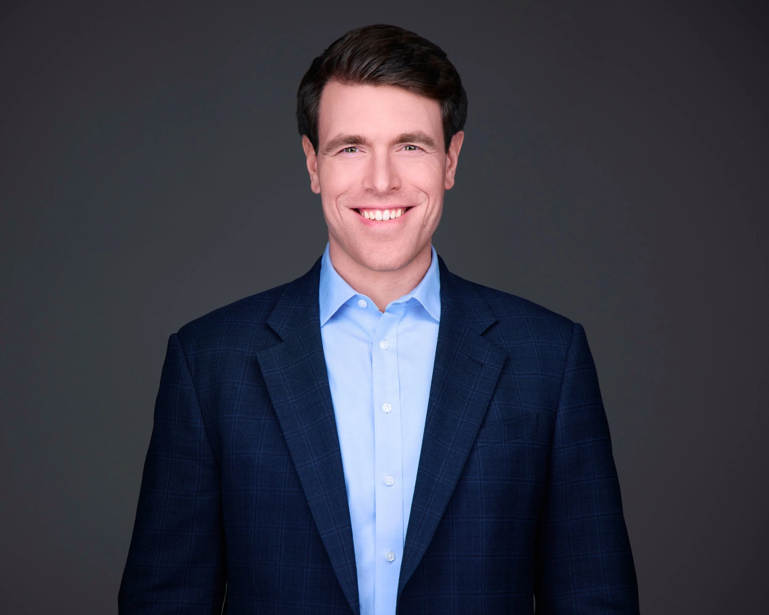 A young man with dark brown hair, wearing a navy blazer and light blue shirt, smiling at the camera against a dark gray background.