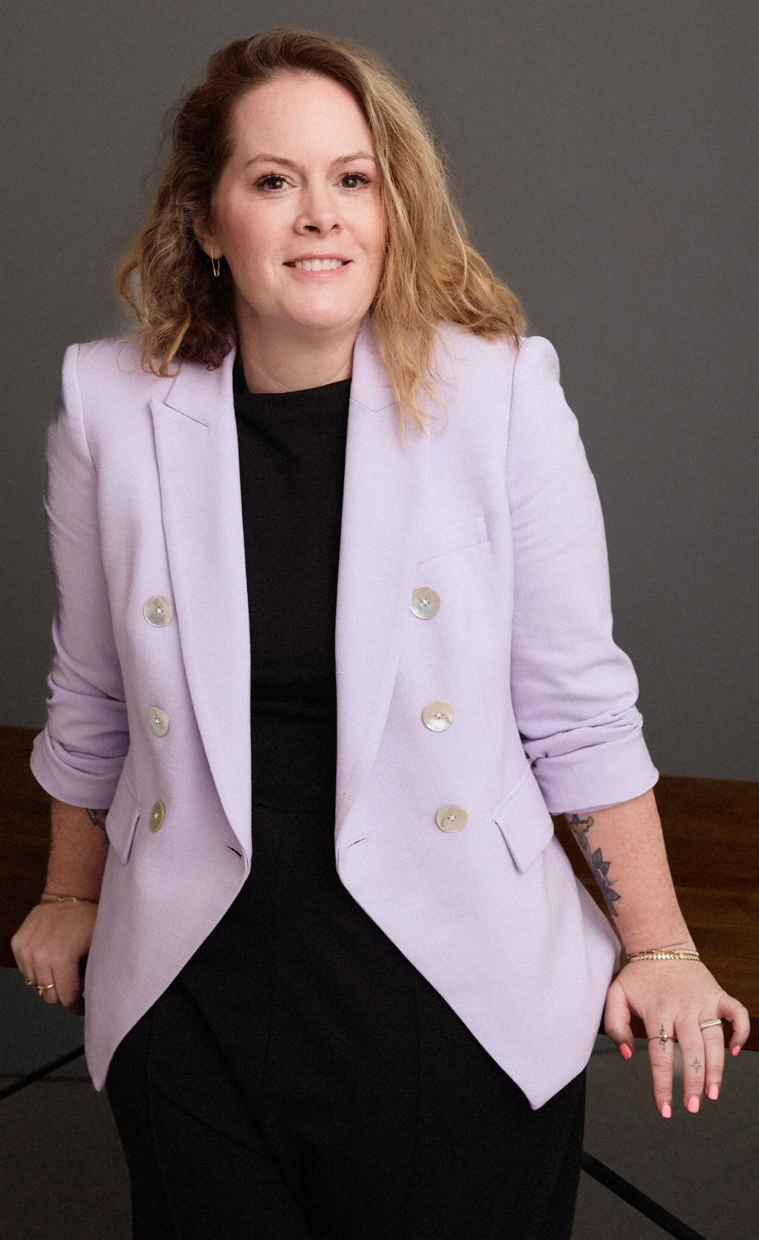 A woman with shoulder-length curly red hair, wearing a lavender blazer with gold buttons and a black top, smiling at the camera against a gray background.