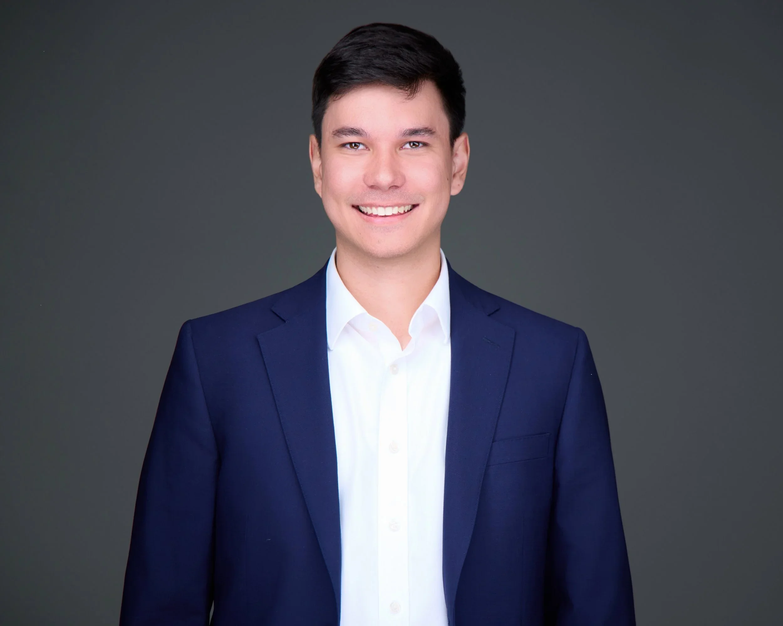 Professional portrait of a young man in a dark blue suit and white shirt, smiling against a dark background.