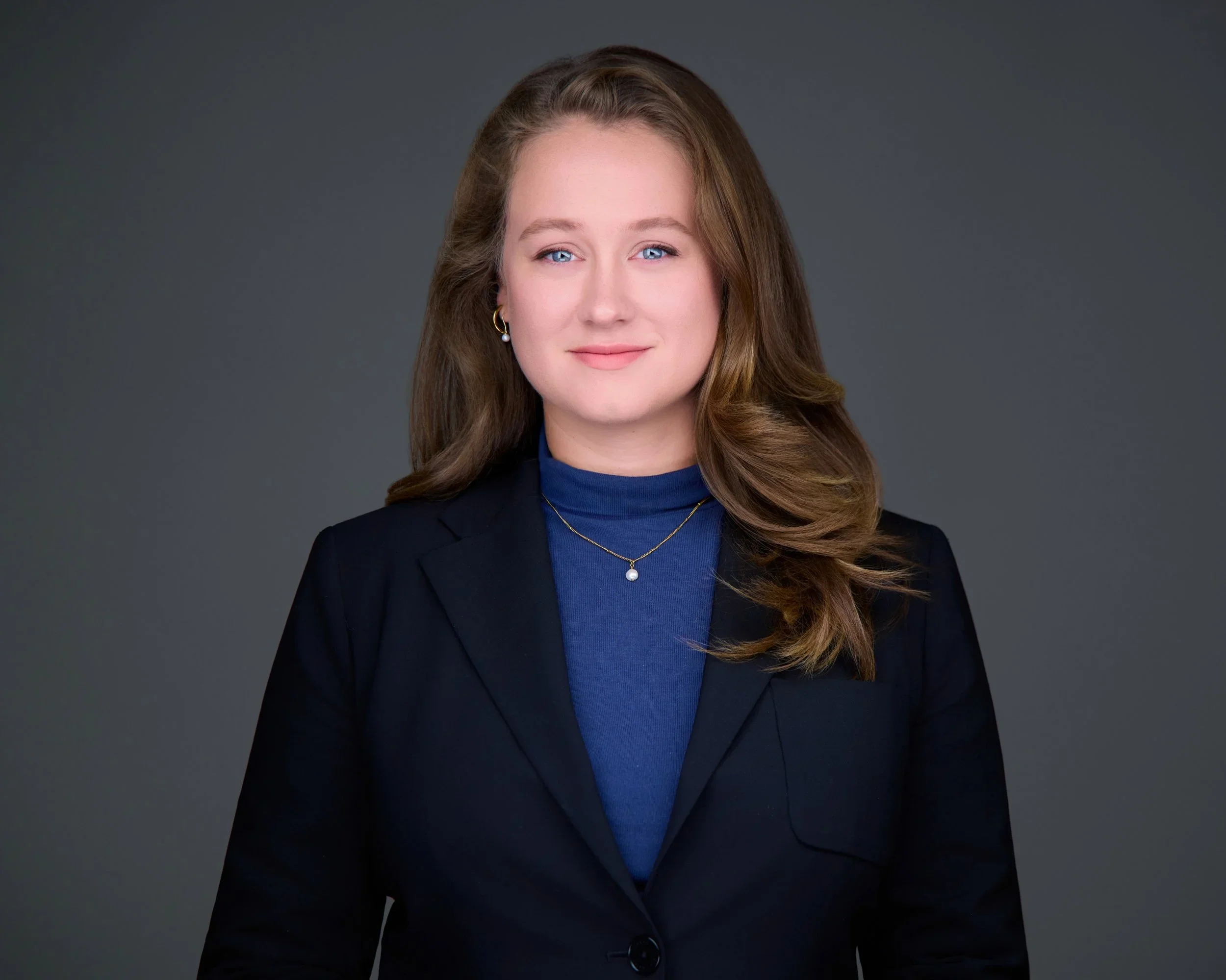 Professional woman with long brown hair, wearing a dark blazer over a blue top, gold jewelry, posing against a gray background.