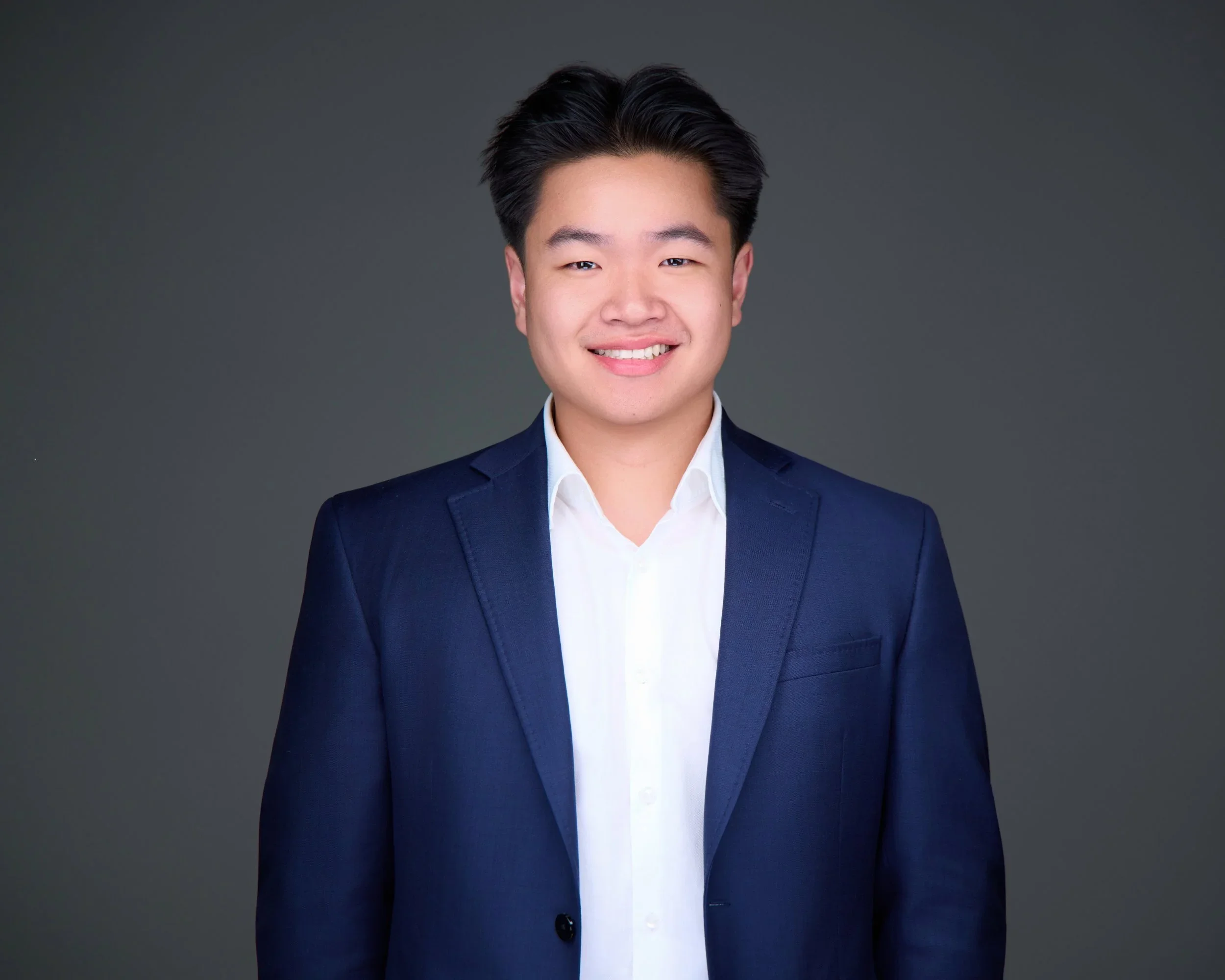 Young man in navy blazer and white shirt smiling against a dark gray background.