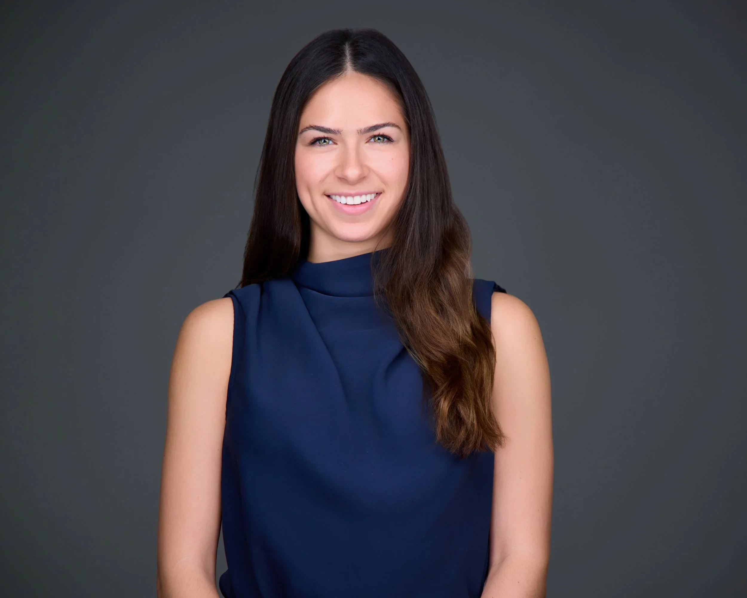 A woman with long dark brown hair smiling, wearing a sleeveless navy blue top, standing against a dark gray background.