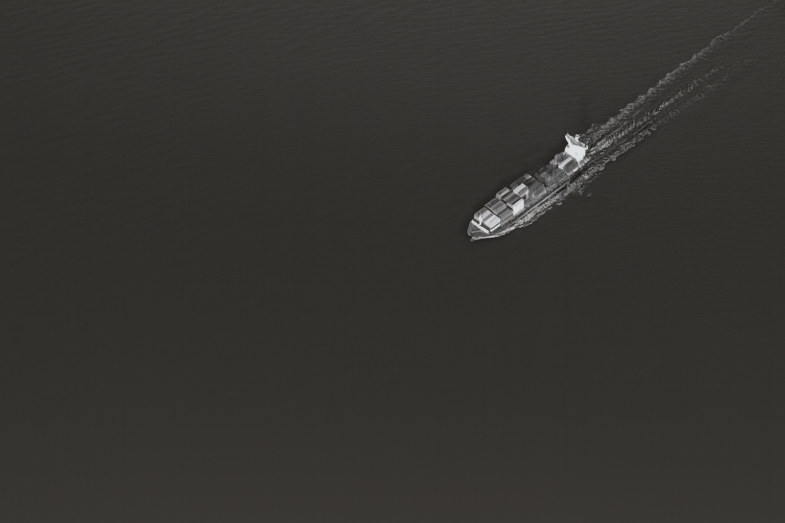 Aerial view of a cargo ship sailing across dark water, leaving a wake behind.