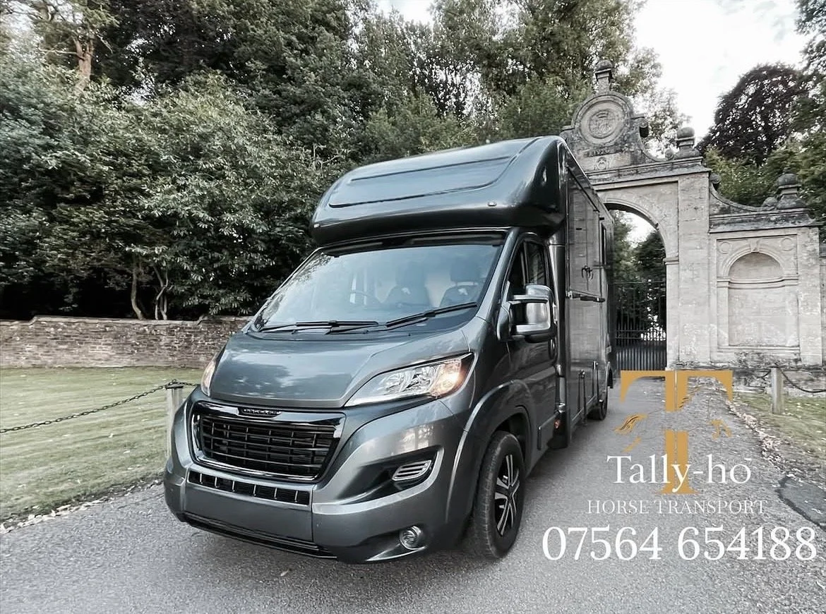 A dark grey horse transport truck parked on a gravel driveway near a stone archway entrance surrounded by trees. Horse transporter near me 