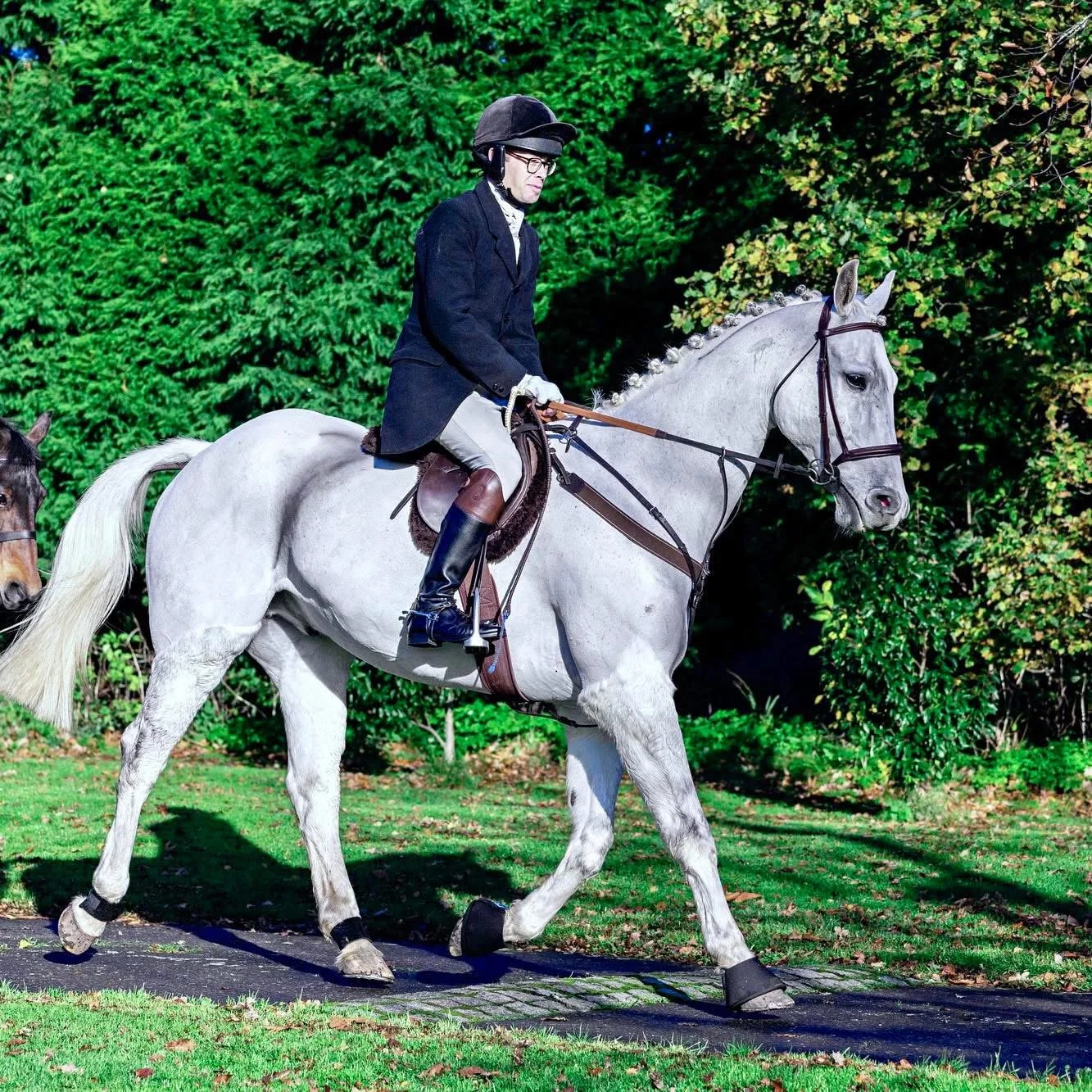 A man wearing a black riding helmet, glasses, a dark blazer, beige riding pants, and tall riding boots riding a white horse on a path outdoors with green foliage in the background.