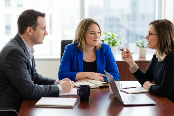 Three professionals engaged in a discussion during a meeting in an office with large windows and plants.