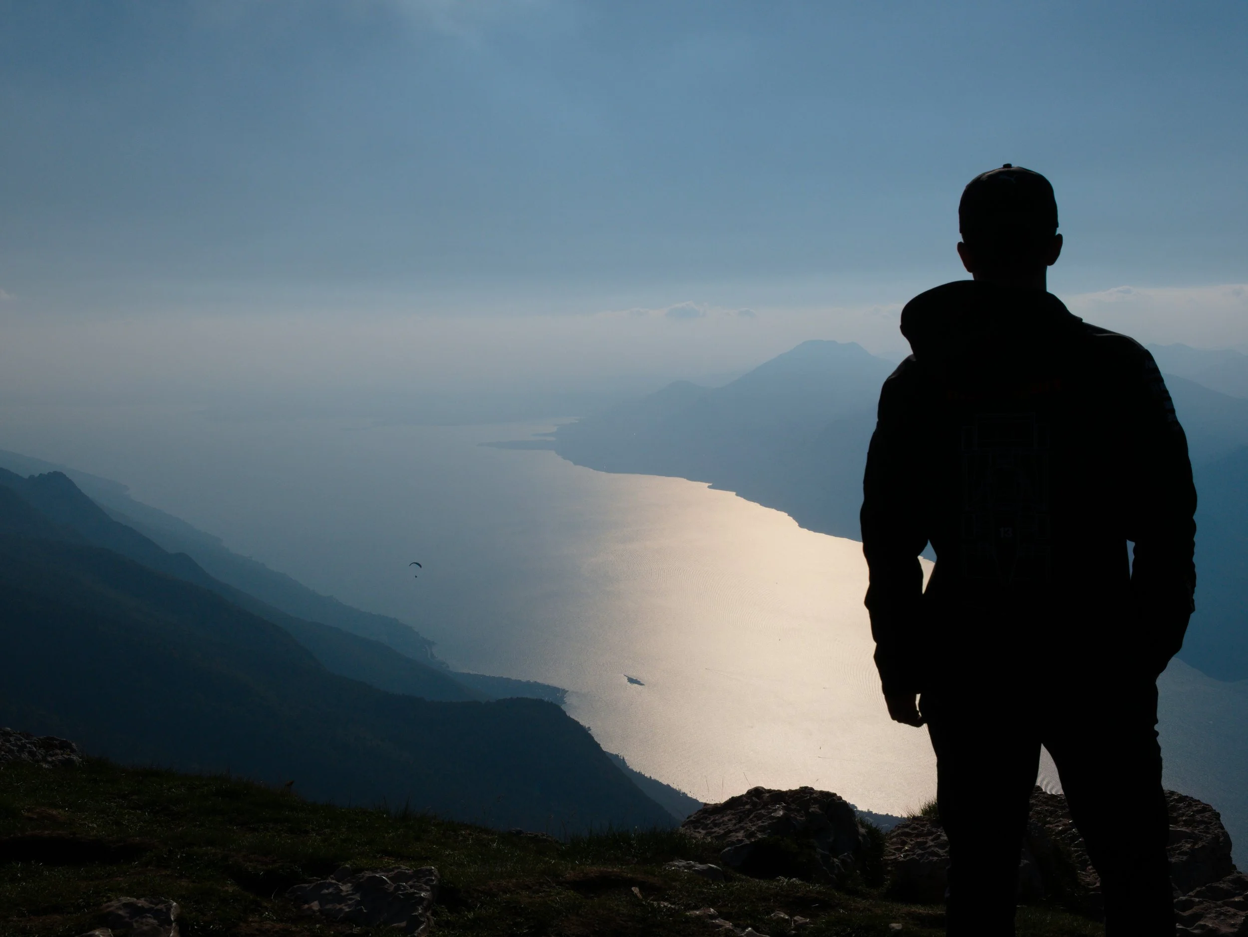 Ein Mann steht mit dem Rücken zur Kamera und blickt auf den Gardasee, umgeben von Bergen, während die Sonne das Wasser reflektiert.