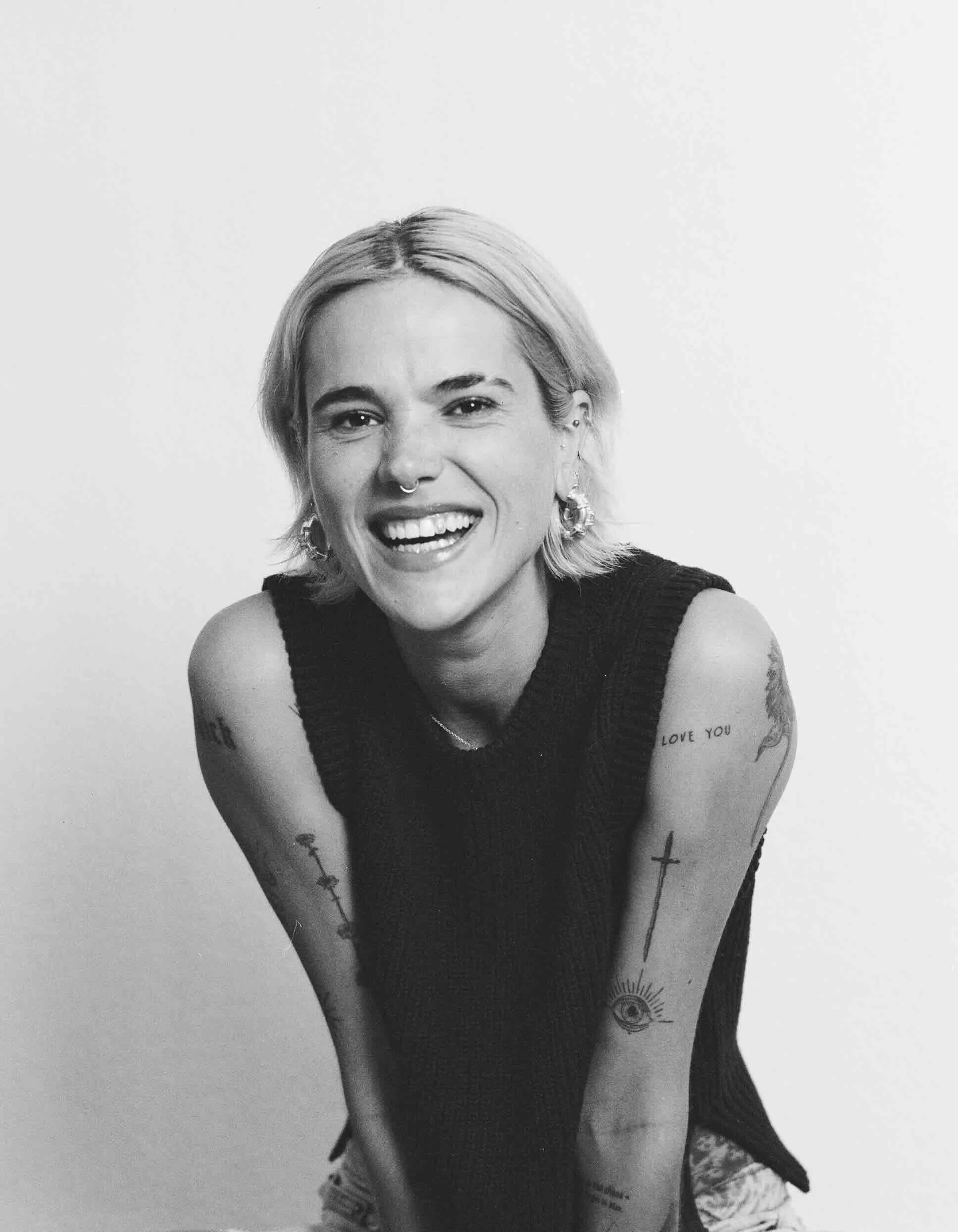Black and white photo of a smiling woman with short blonde hair, wearing a sleeveless black top, earrings, and tattoos on her arms.