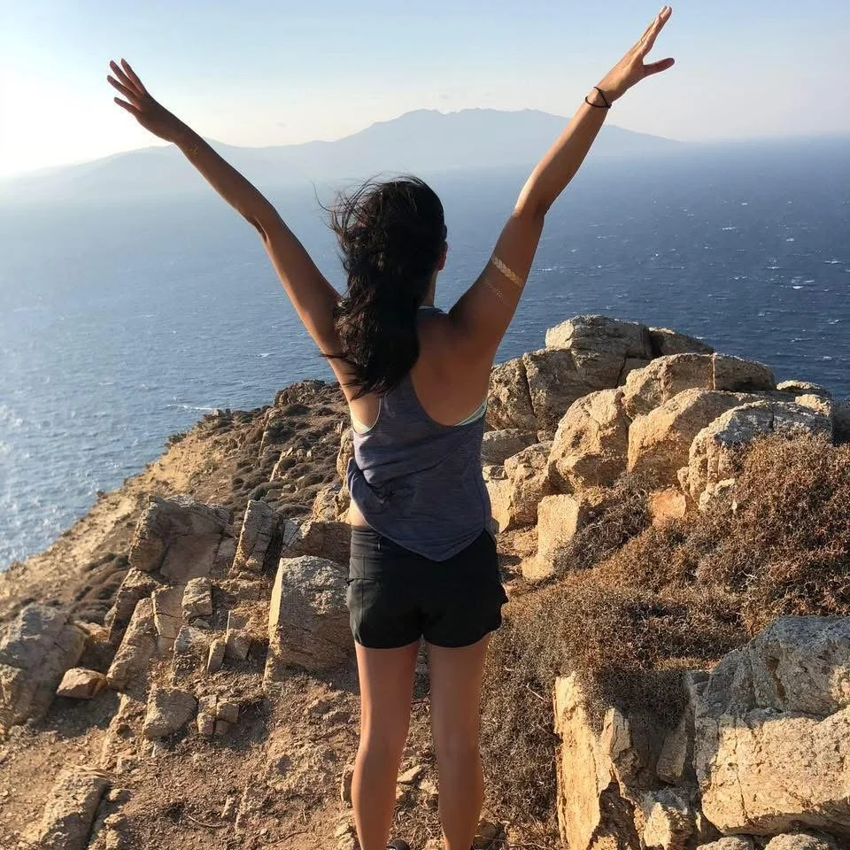 A woman stands with her arms raised in front of a scenic ocean view from a rocky cliff.