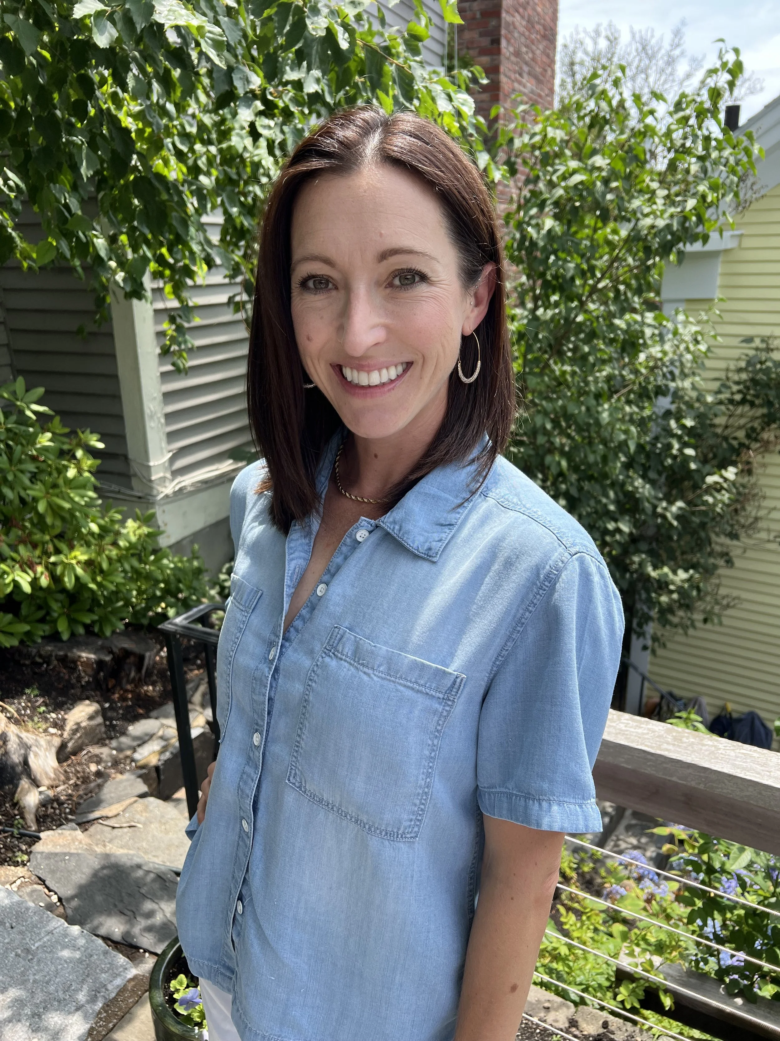 A woman with shoulder-length brown hair smiling outdoors, wearing a light blue denim shirt, in front of green bushes and trees.