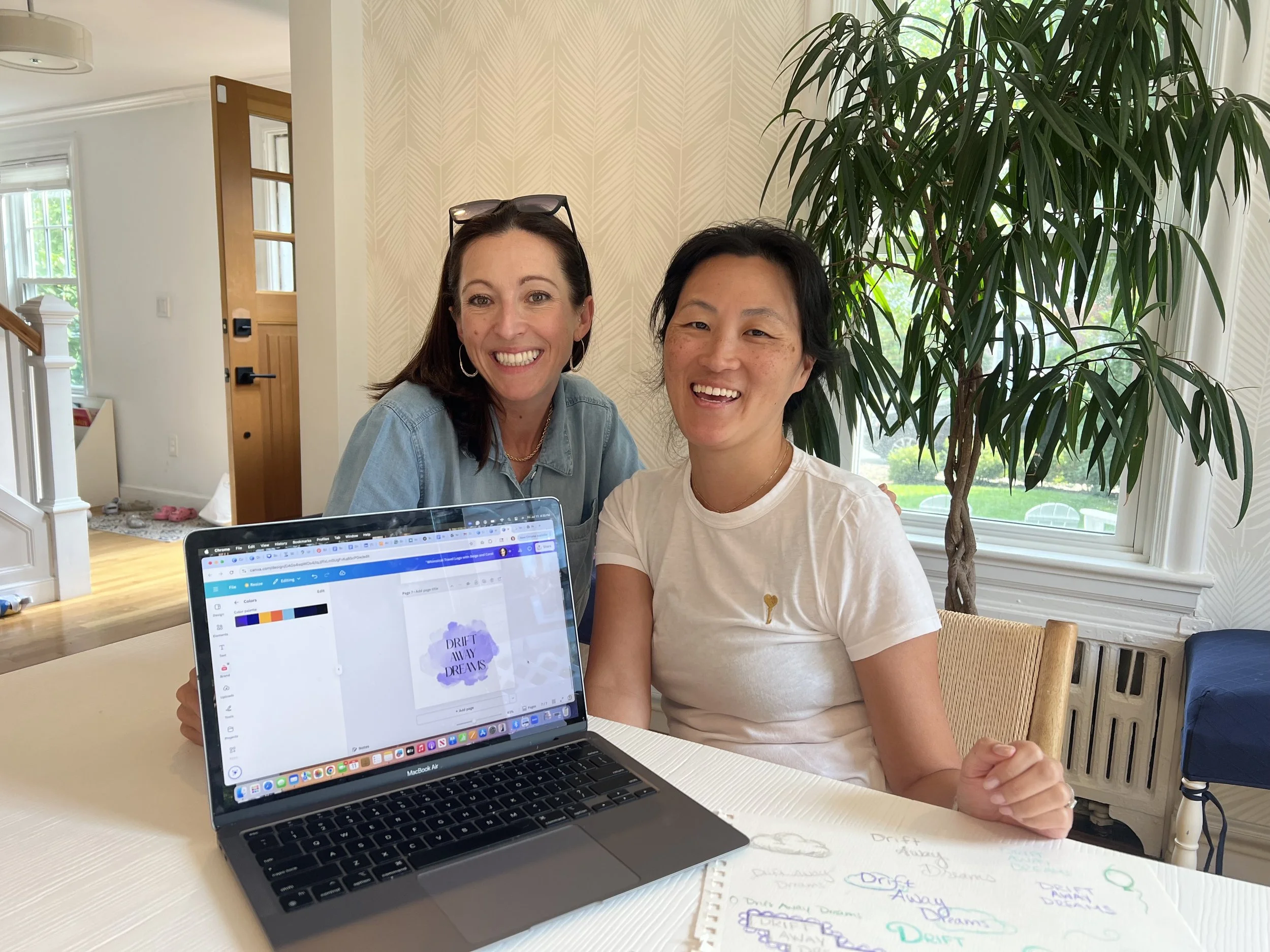 Two women sitting at a table with a laptop and a notebook, smiling at the camera in a bright room with a large window and a tall potted plant.