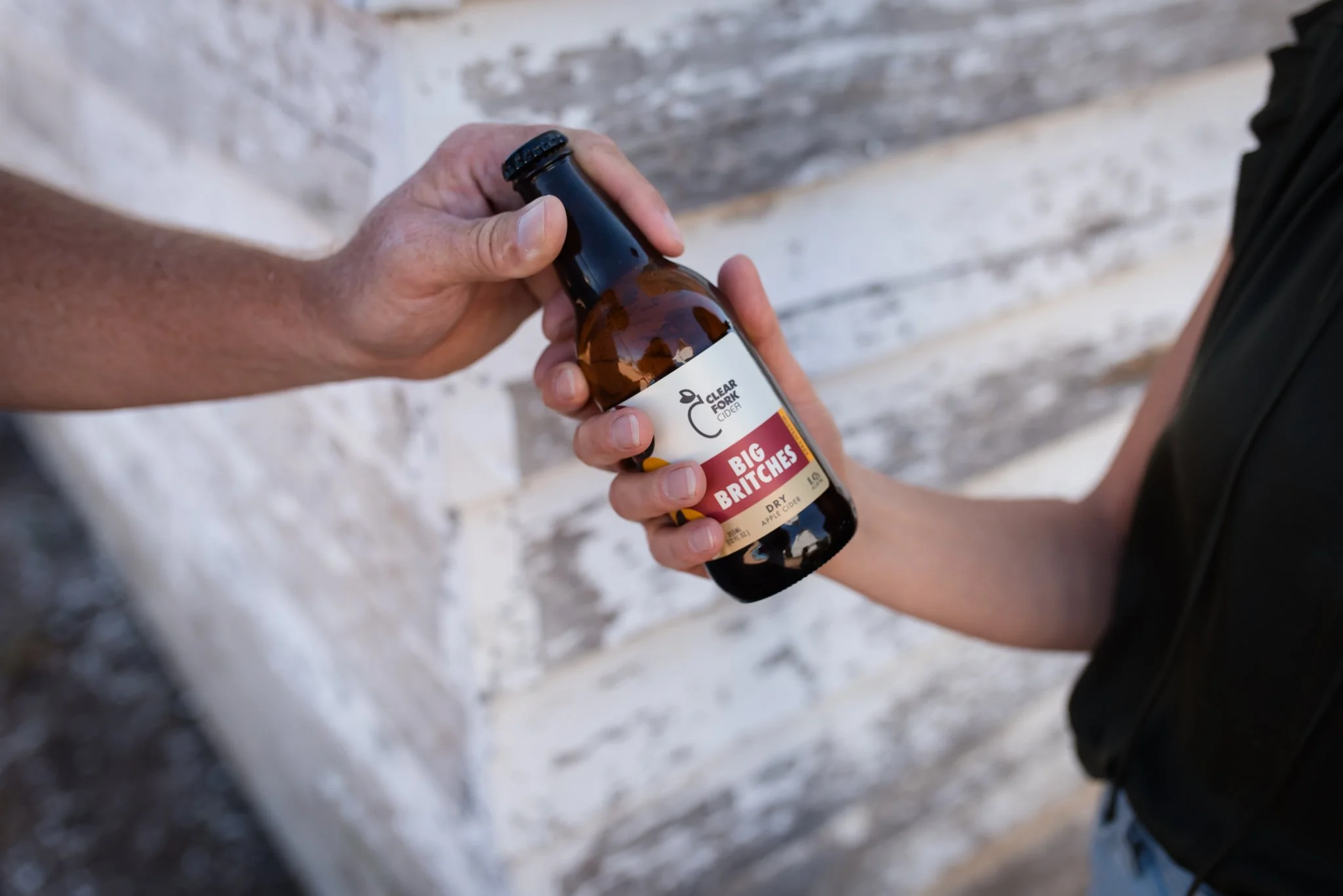A person handing a bottle of Clear Fork Cider to another person in front of a wooden wall.