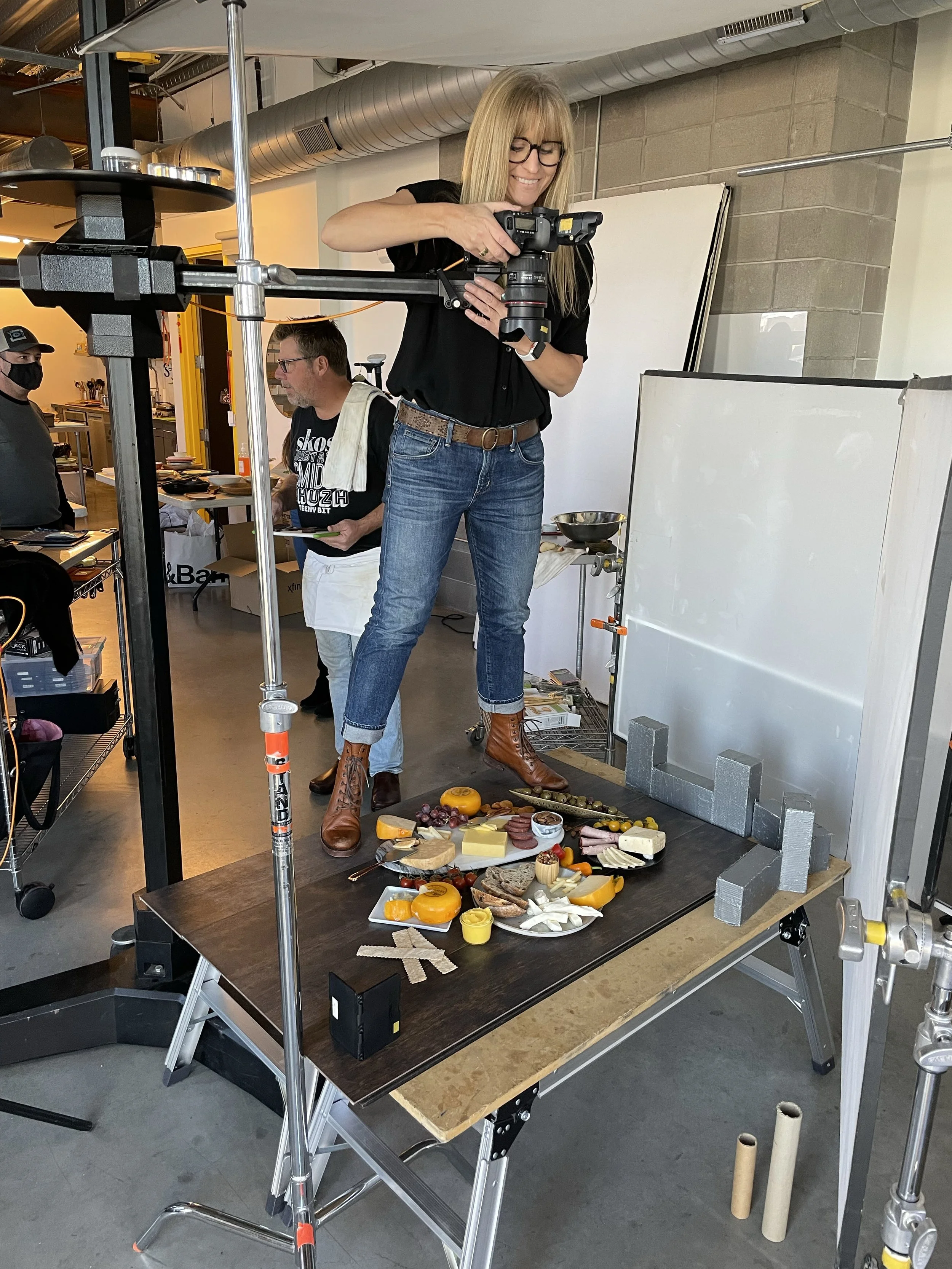 A woman stands on a platform to arrange or style food props on a table, which includes cheese, meats, fruits, and decorative items, in an indoor setting. She is holding a camera, smiling, and wearing glasses, a black shirt, blue jeans, and brown boots.