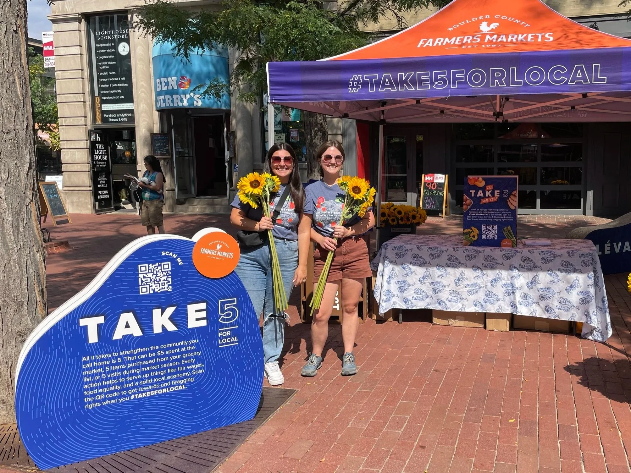 Two women standing outdoors at a community event, holding sunflowers, in front of an informational sign and a table under a purple canopy with the hashtag #TAKE5FORLOCAL, promoting local shopping with a QR code for scanning, neighboring storefronts visible.
