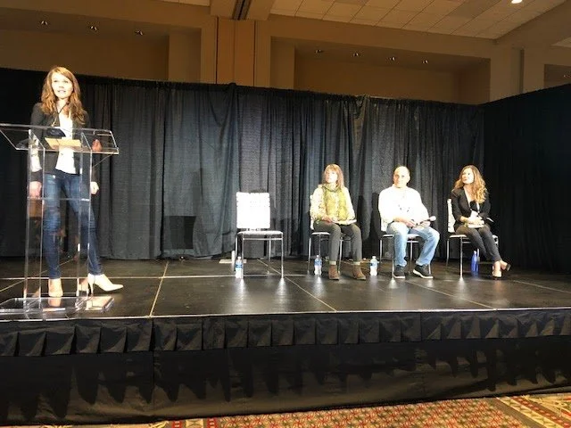 A woman speaking at a podium on a stage with three people seated behind her, captured during a panel or presentation event in a conference room.