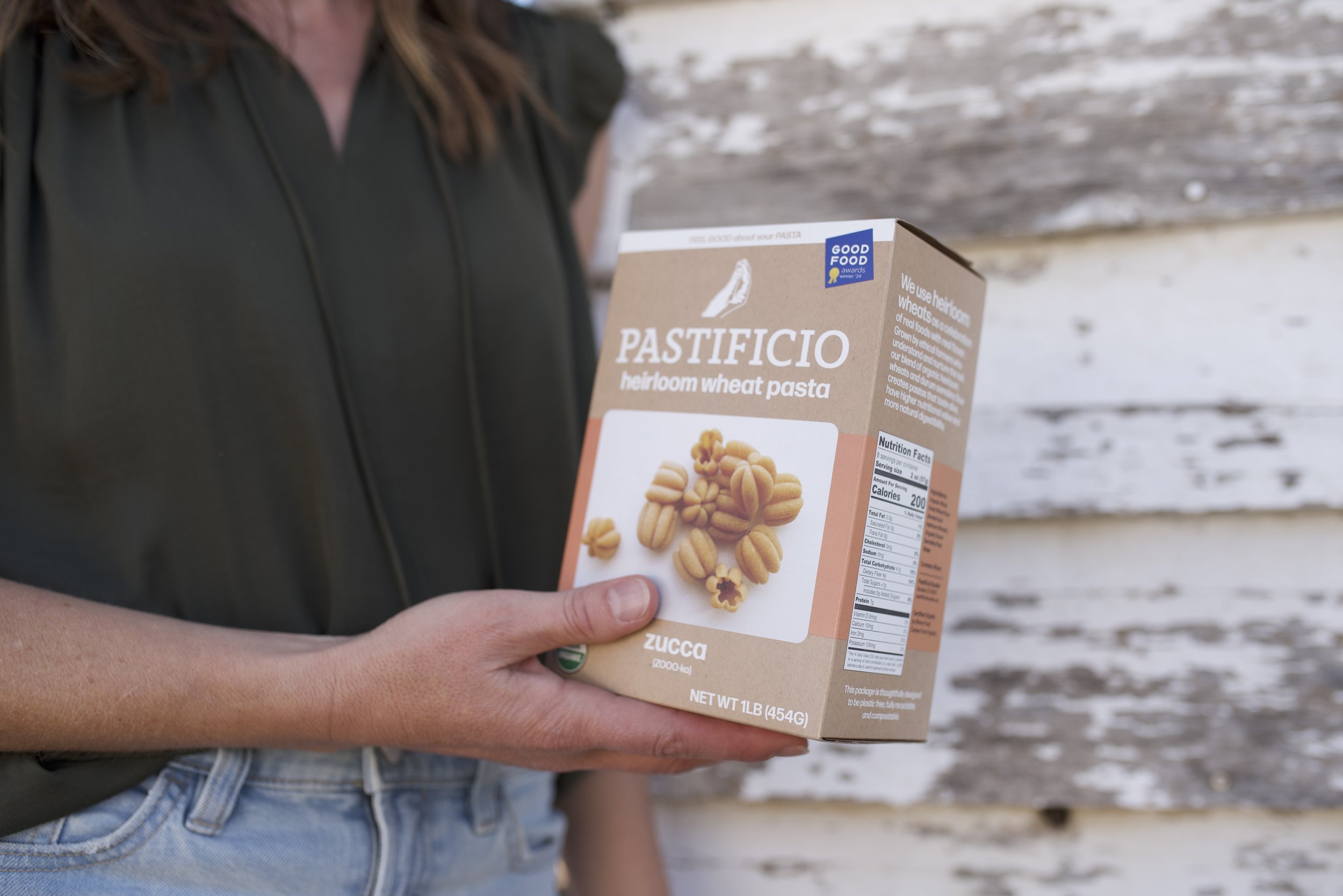Person holding a box of Pastificio heirloom wheat pasta in front of a weathered wooden wall.