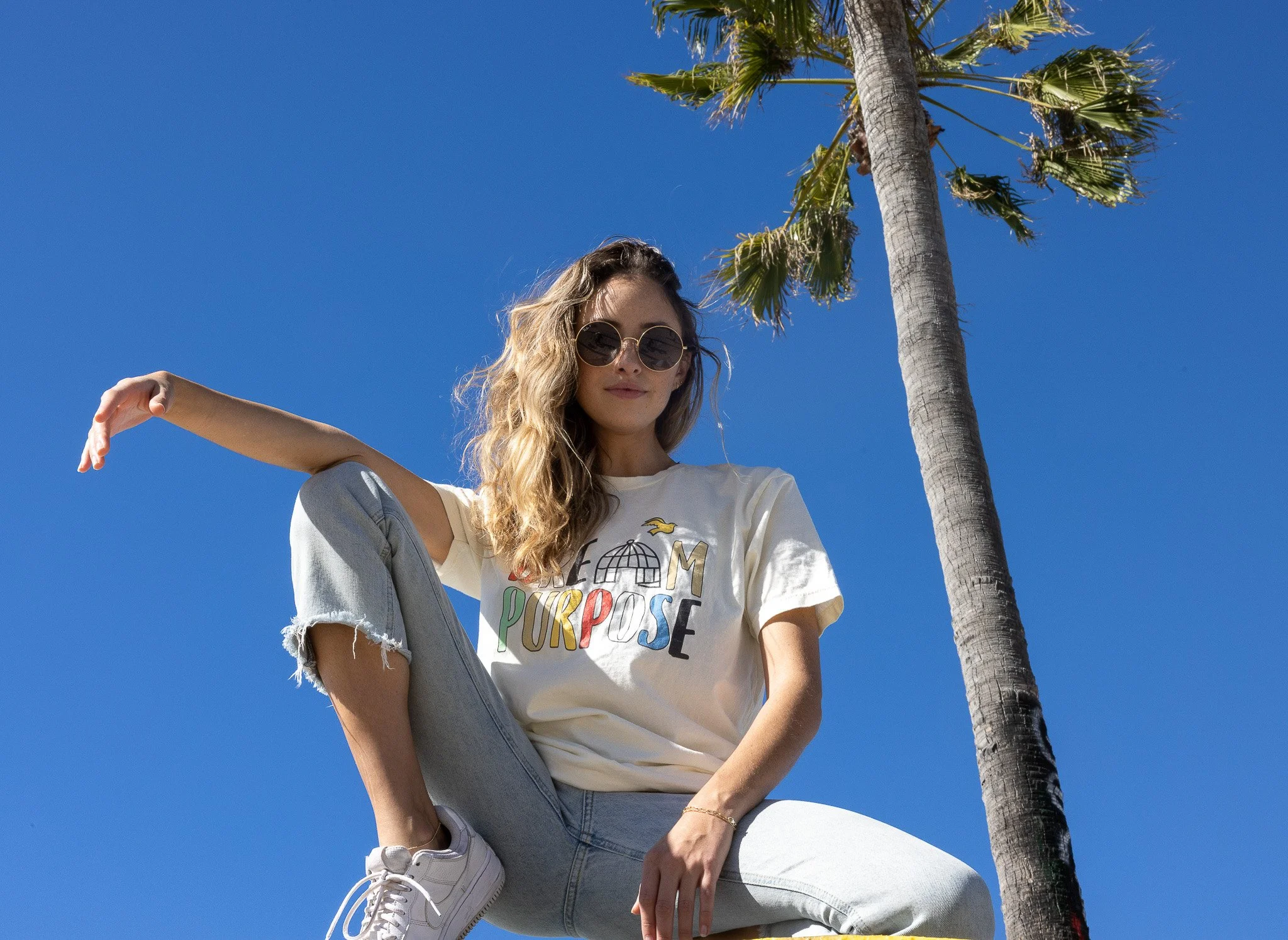 A young woman with curly hair wearing sunglasses, a cream T-shirt with colorful lettering, and ripped jeans, posing outdoors next to a tall palm tree against a clear blue sky.