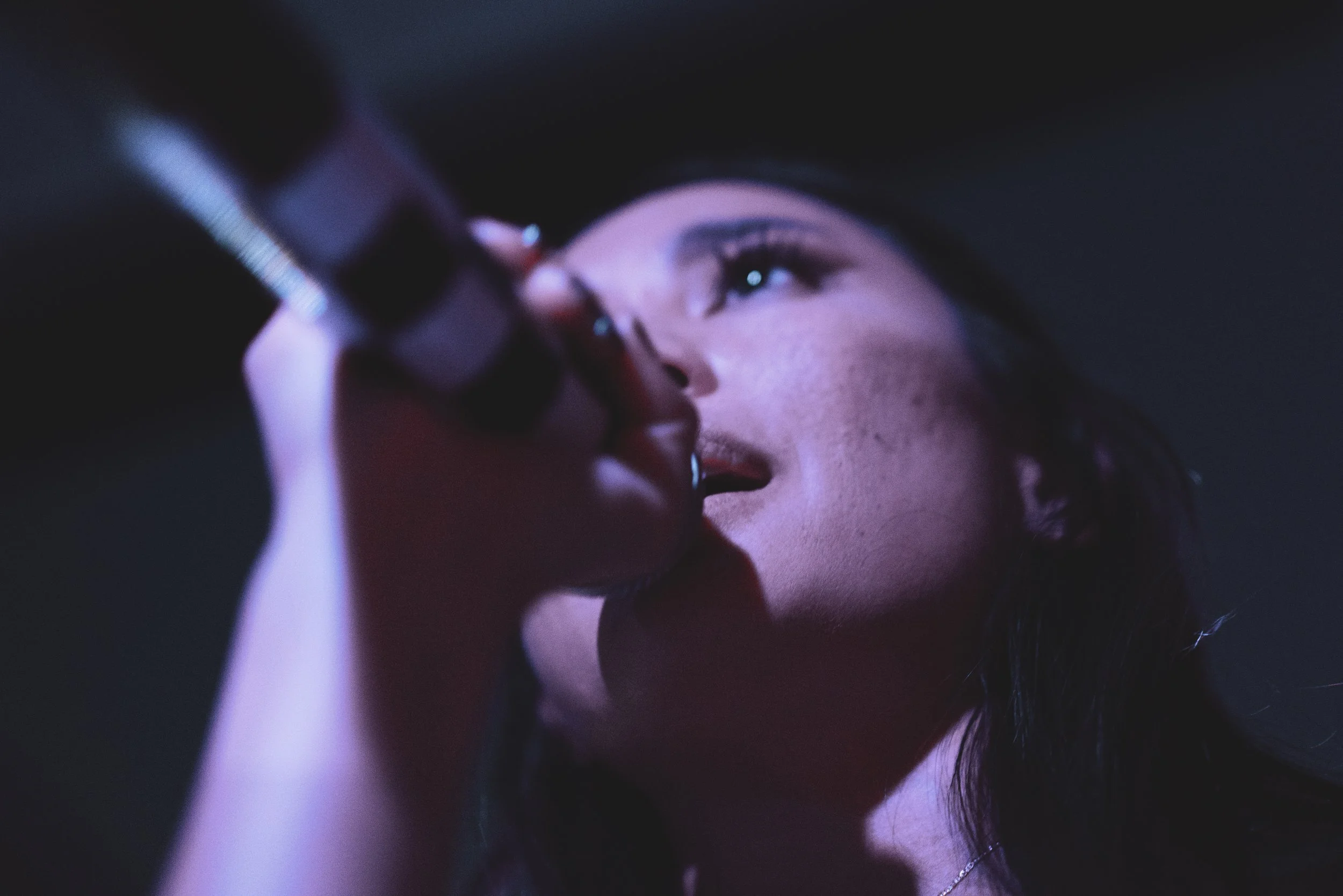 A woman applies lipstick in a dimly lit setting with a purple hue, looking slightly upward with her eyes open.