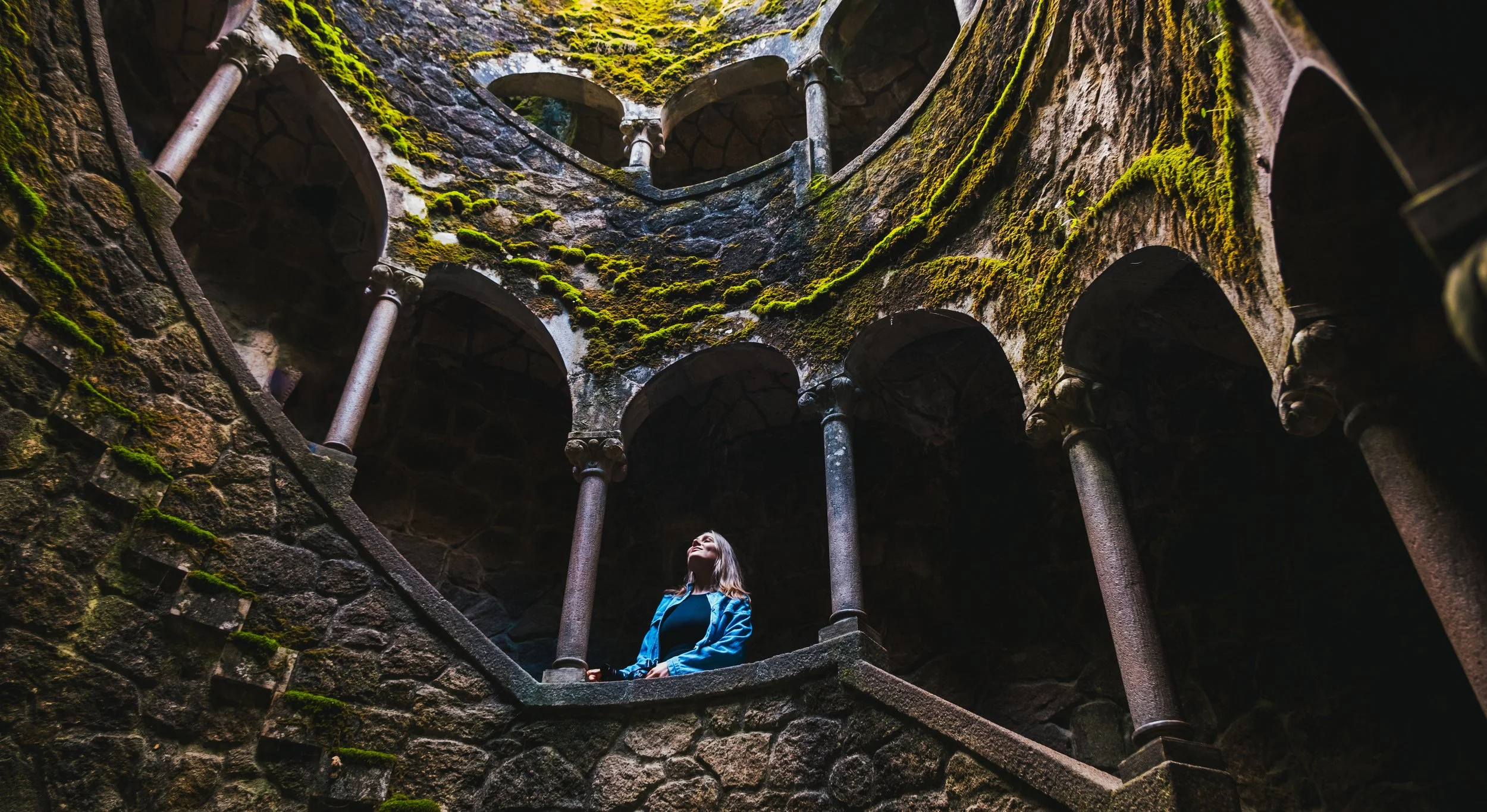 A woman in a blue jacket standing inside the moss-covered ruins of a stone building, looking upward.