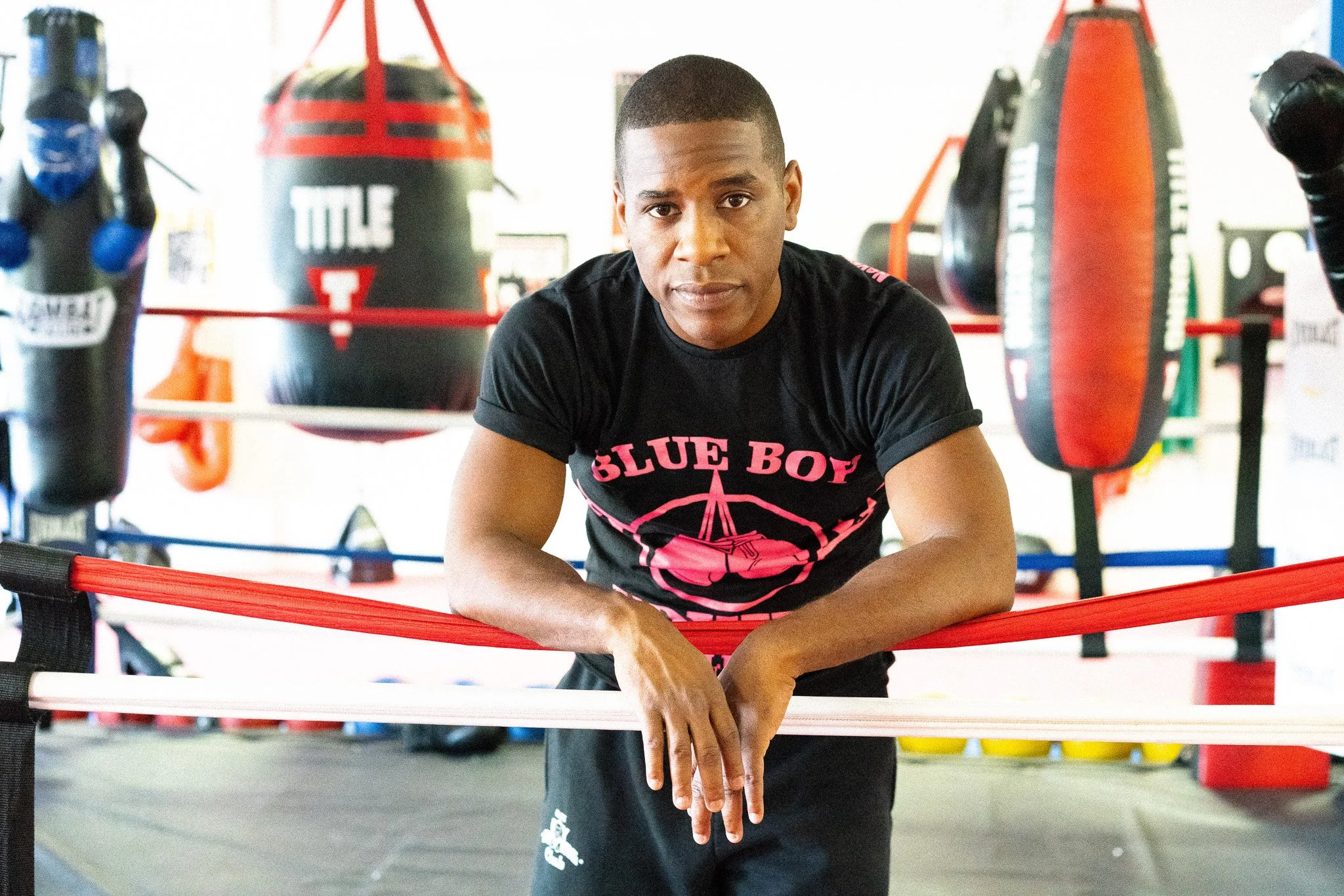 A man in a black t-shirt with pink text and graphics, leaning on a boxing ring's red and white ropes in a boxing gym.