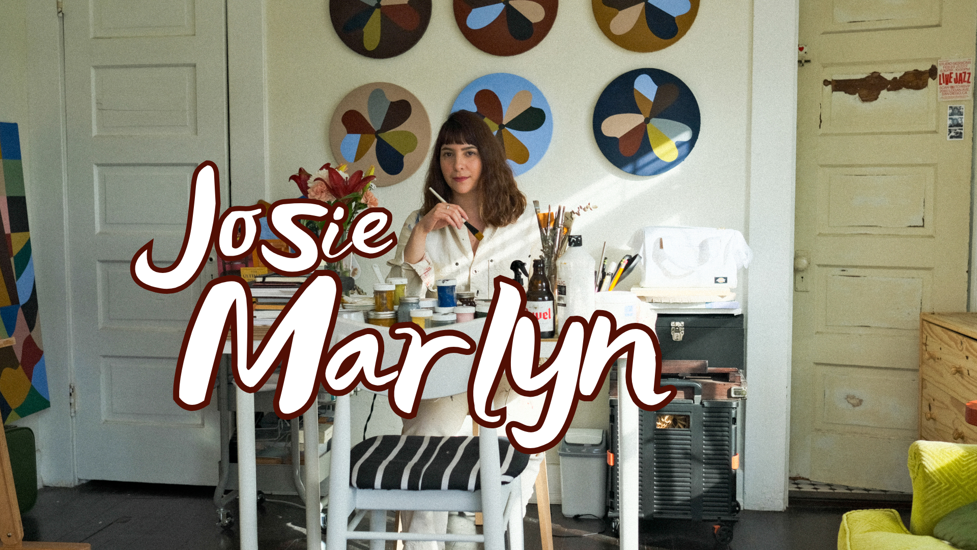 A woman with curly brown hair and a white shirt sitting at an art studio table with various art supplies, including paints, brushes, and notebooks. Behind her, colorful circular abstract paintings are hung on the wall. The text 'Josie Marlyn' is written across the lower part of the image.