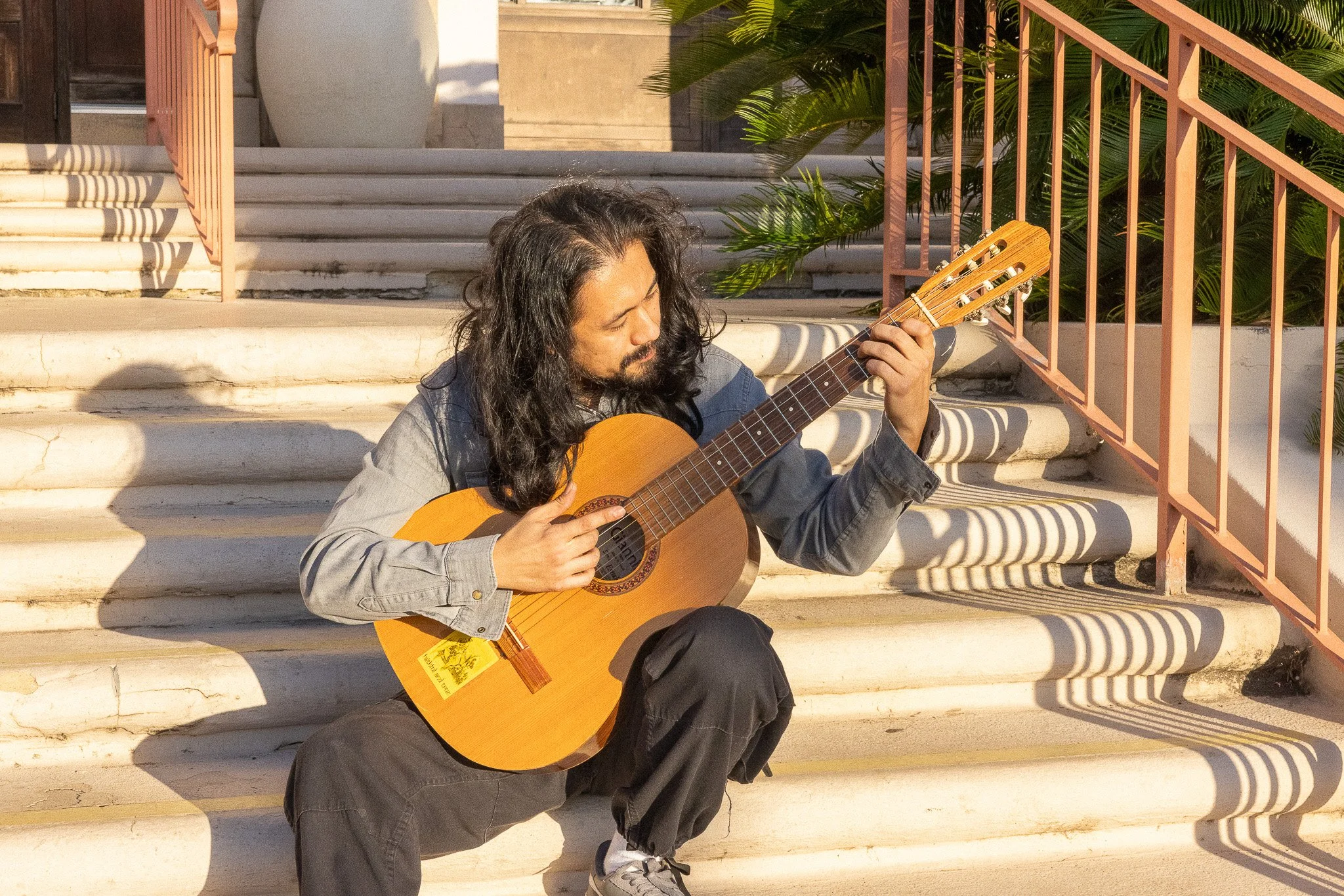A man with long curly hair and a beard playing an acoustic guitar while sitting on steps outside.