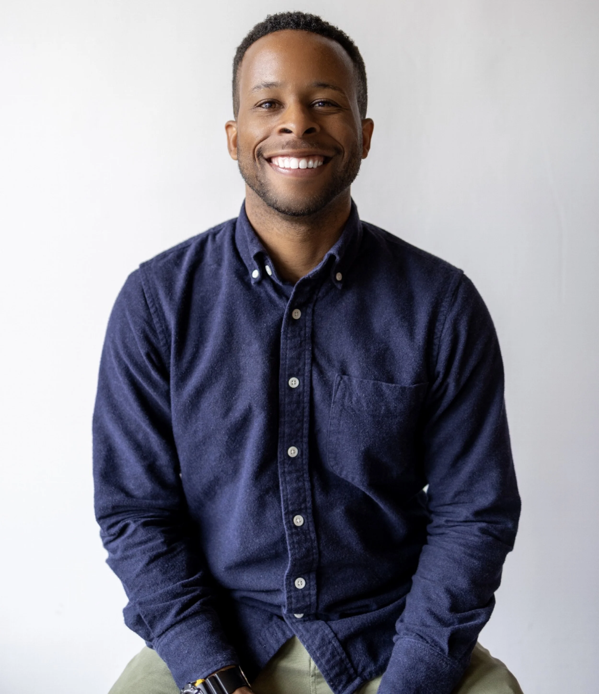 A smiling man with short dark hair, wearing a navy button-down shirt and khaki pants, sitting against a plain white background.