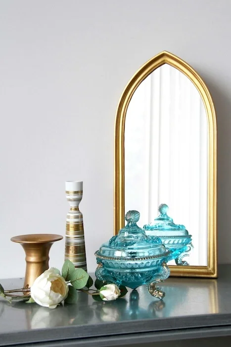 A dresser with gold vases, a glass blue decorative container, and a gold-trimmed mirror on top of it.