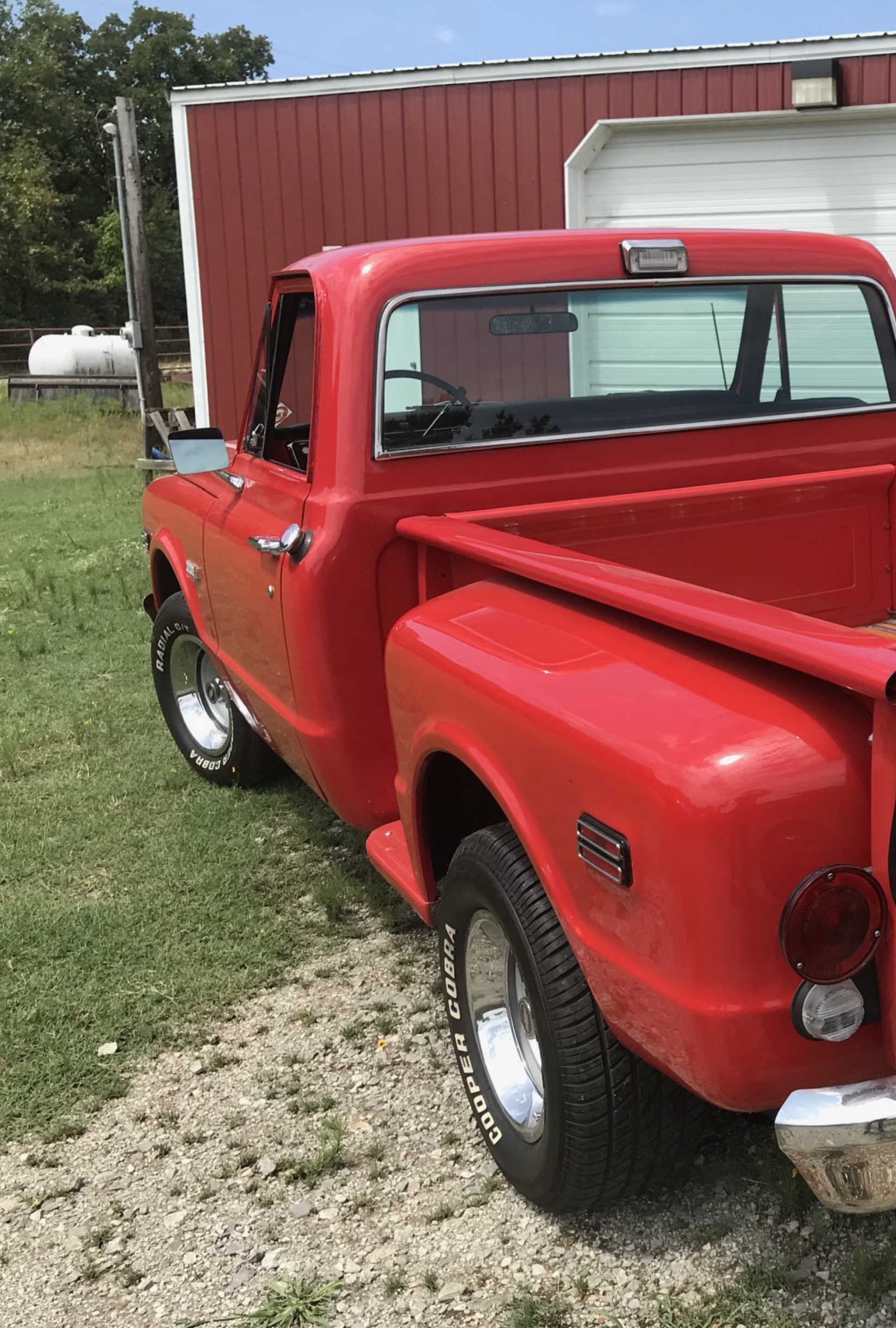 A red vintage pickup truck parked on a grassy area near a red barn with a white garage door.
