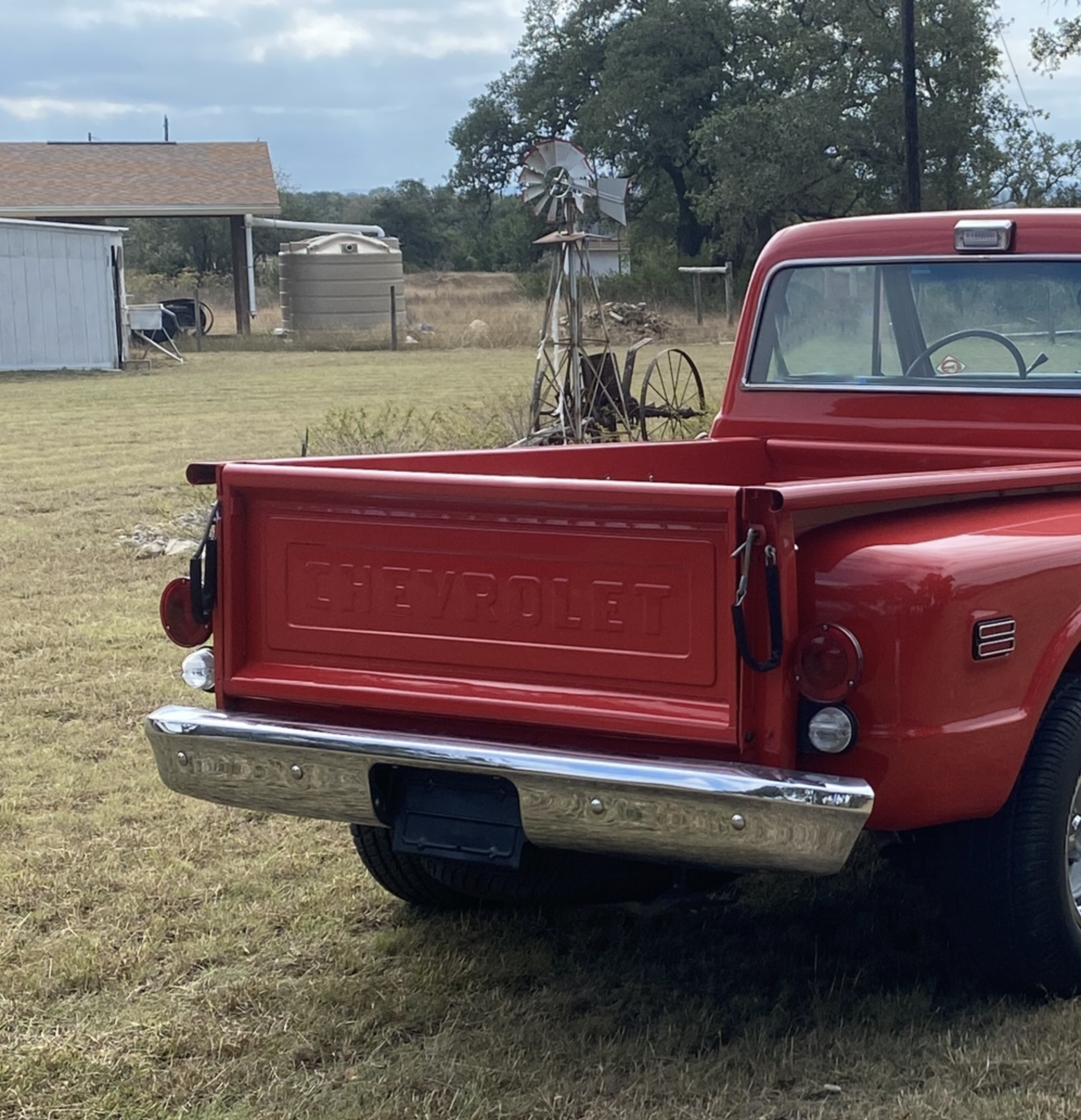 Red Chevrolet pickup truck parked on a grassy field with farm structures and a windmill in the background.