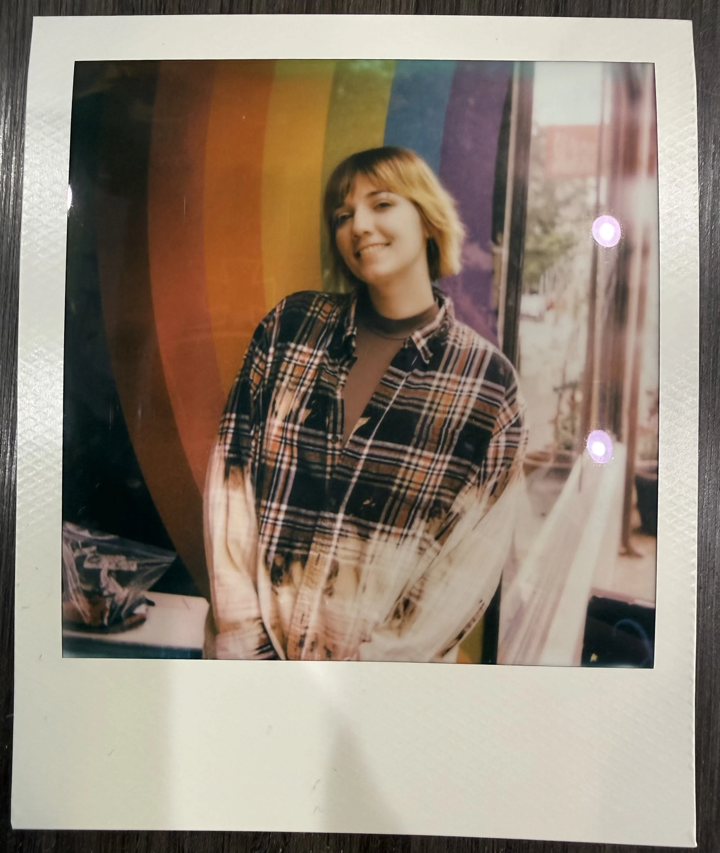 A print of a young woman with shoulder-length blonde hair, wearing a plaid shirt and a dark turtleneck, standing in front of a rainbow-colored background with vertical stripes, smiling slightly at the camera.