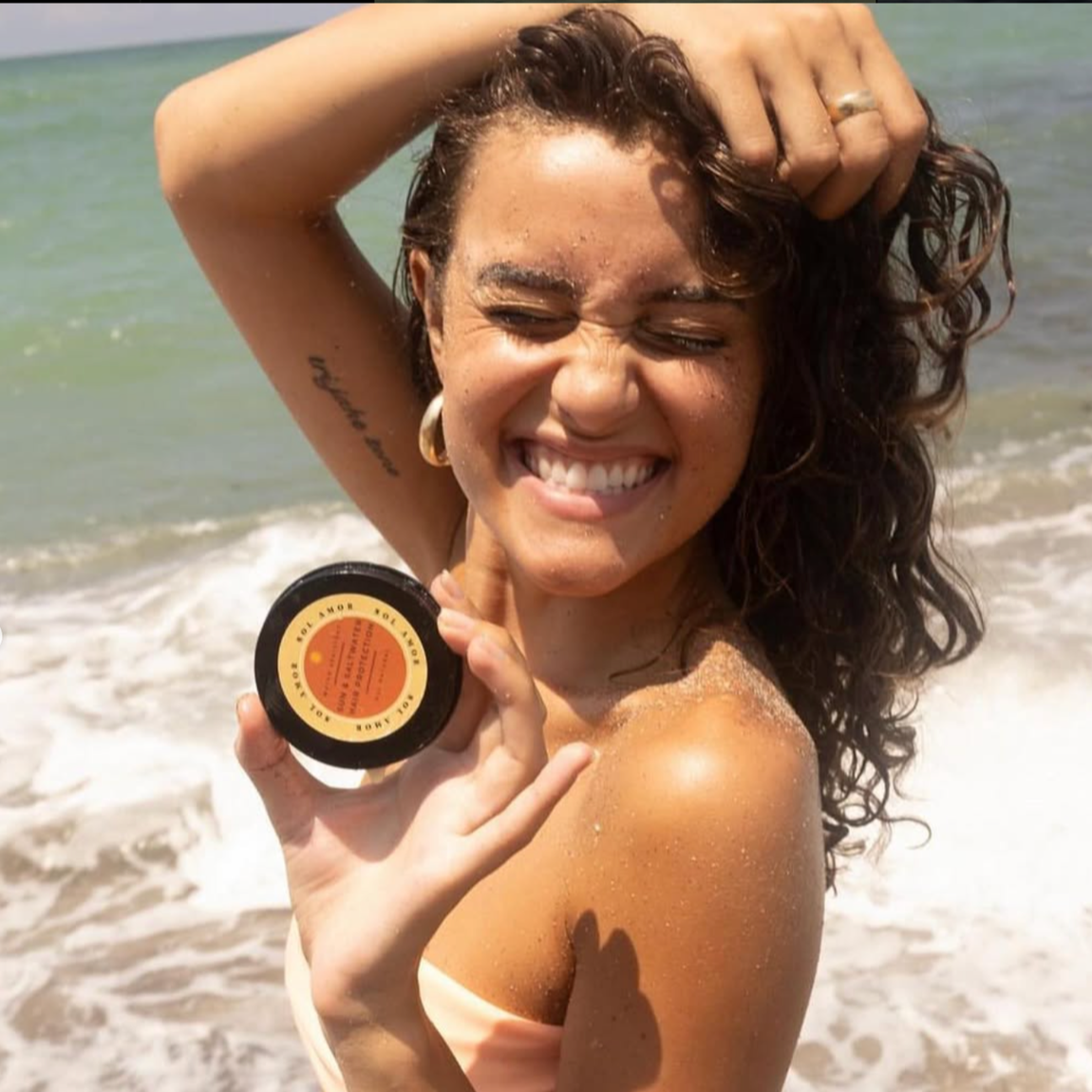 Woman at the beach smiling, holding a small container of skincare or sunscreen, with waves in the background, wearing hoop earrings and a swimsuit.