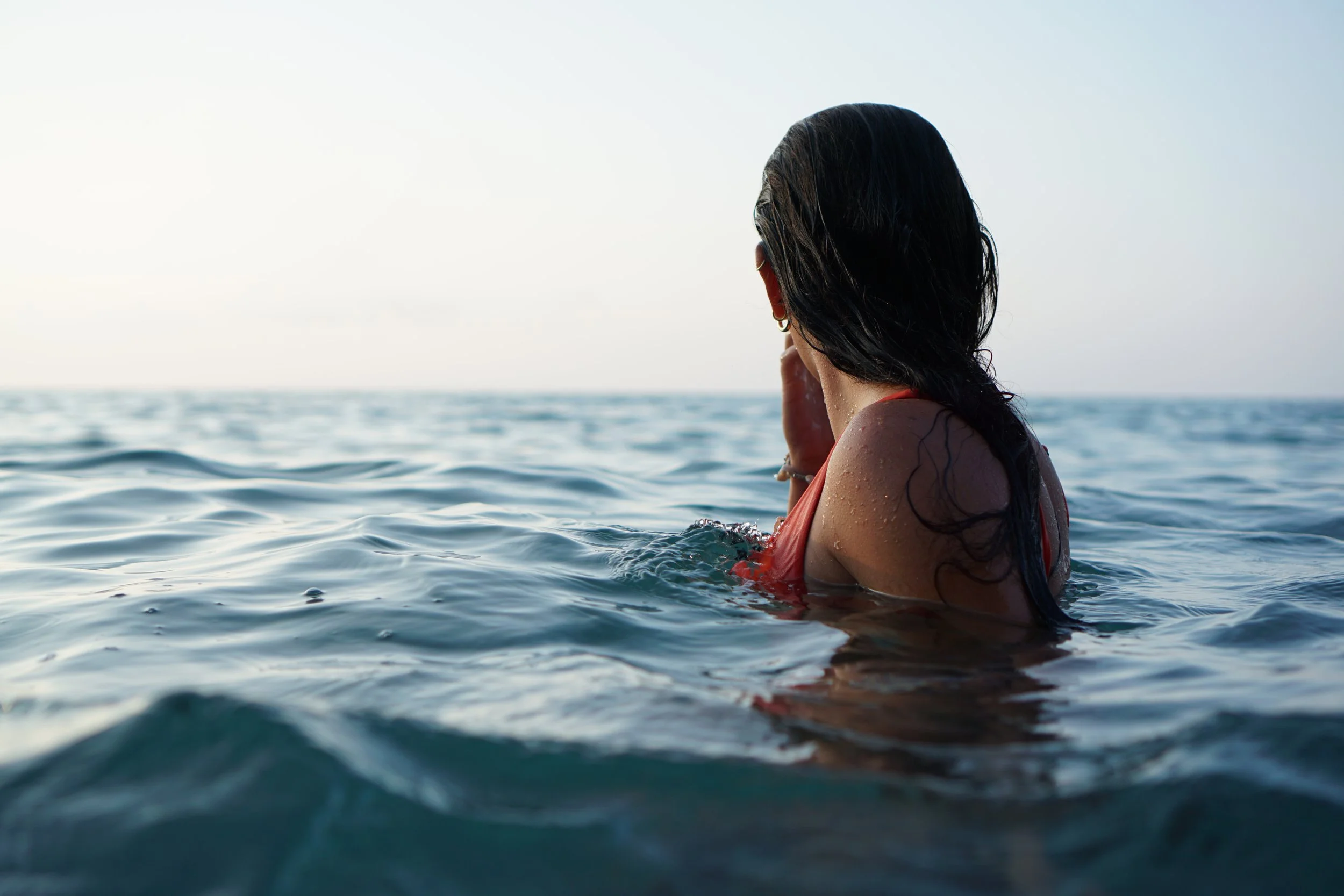 A woman with dark hair in a red swimsuit swimming in the ocean, facing away from the camera, with the sky in the background.