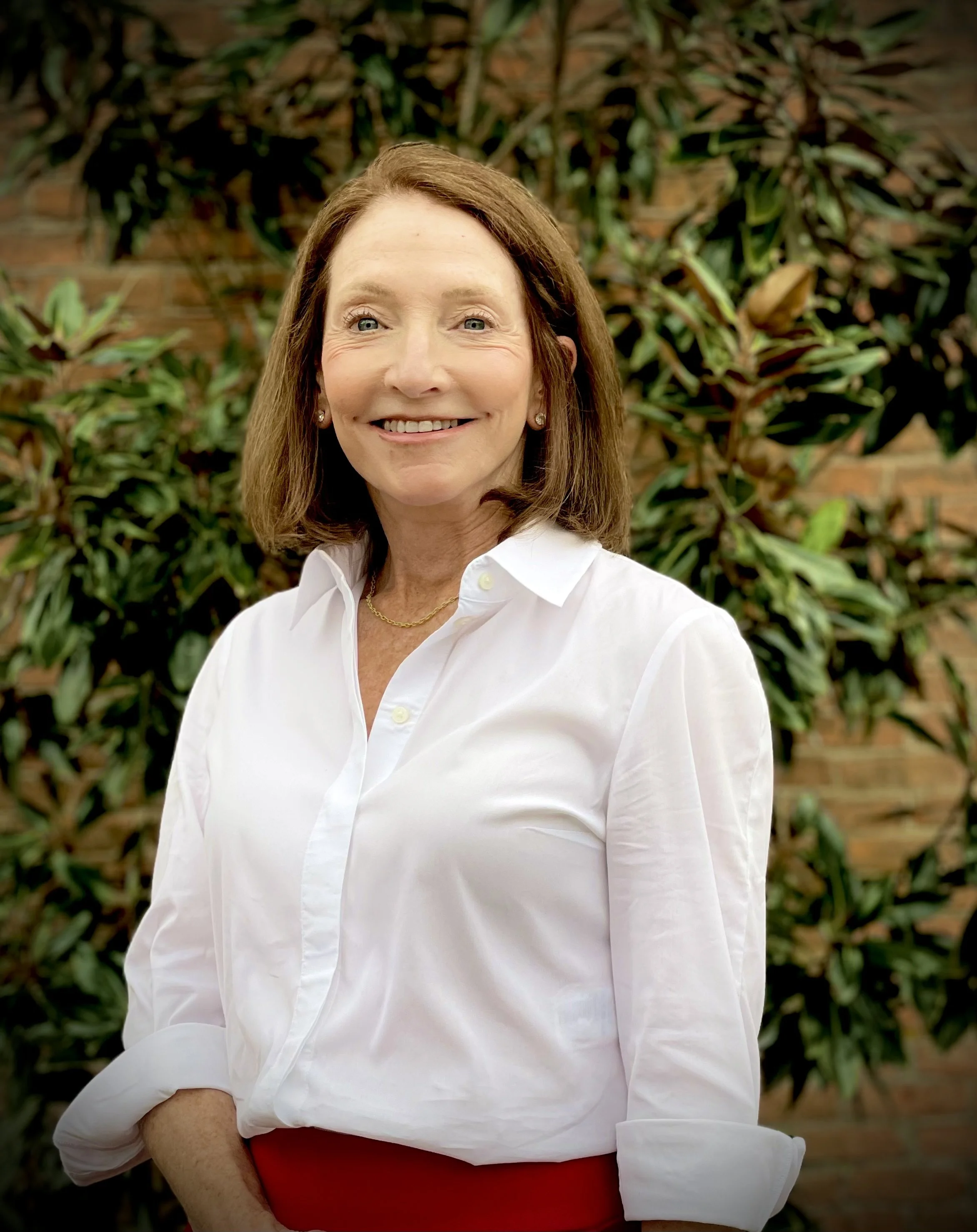 A woman with shoulder-length brown hair, wearing a white blouse and a gold necklace, standing outdoors in front of green leafy plants and a brick wall, smiling at the camera.