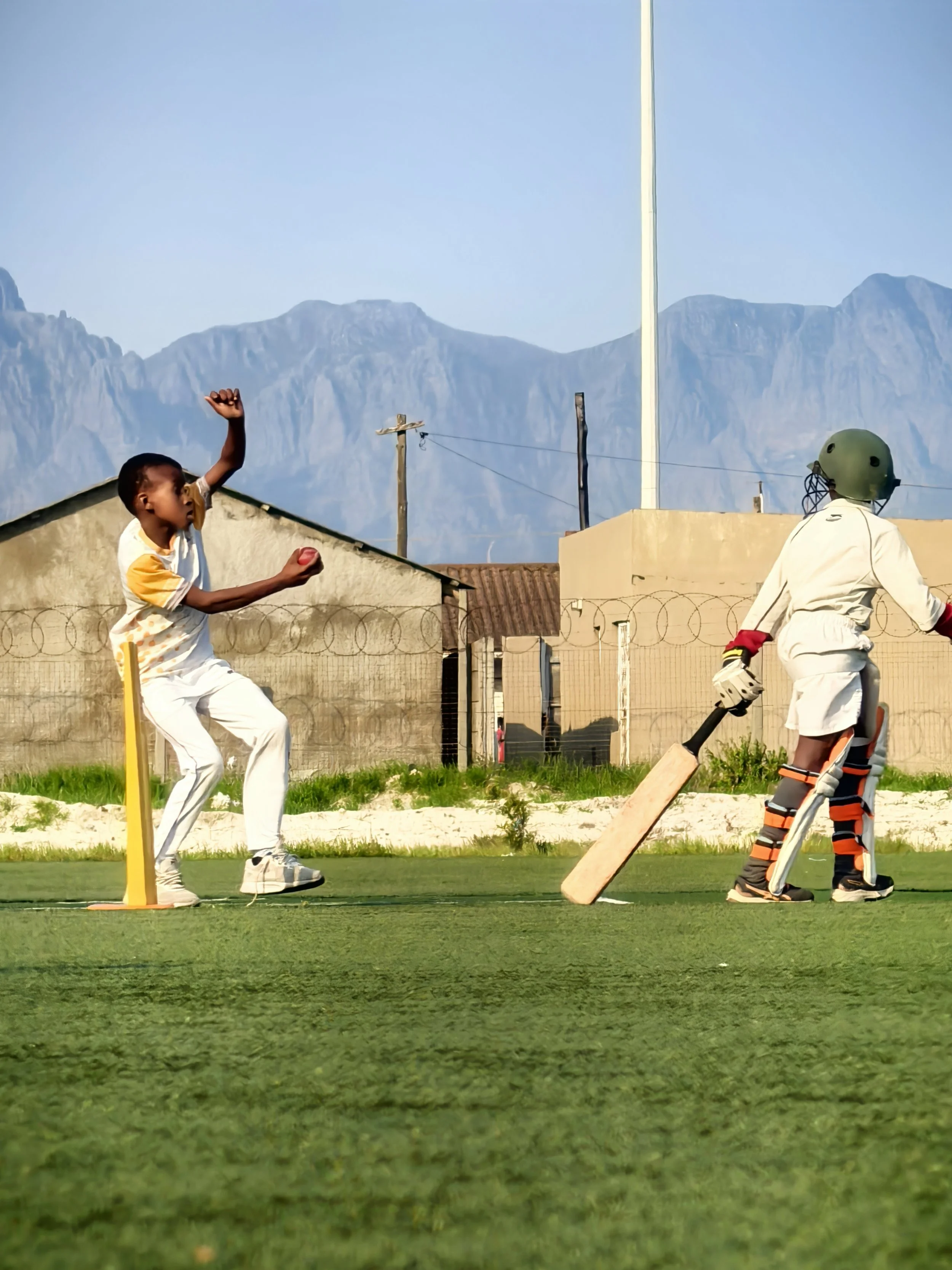 Two children playing cricket outdoors on a grassy field with mountains in the background. One child is preparing to hit the ball, and the other is ready to bowl, wearing a helmet and protective gear.
